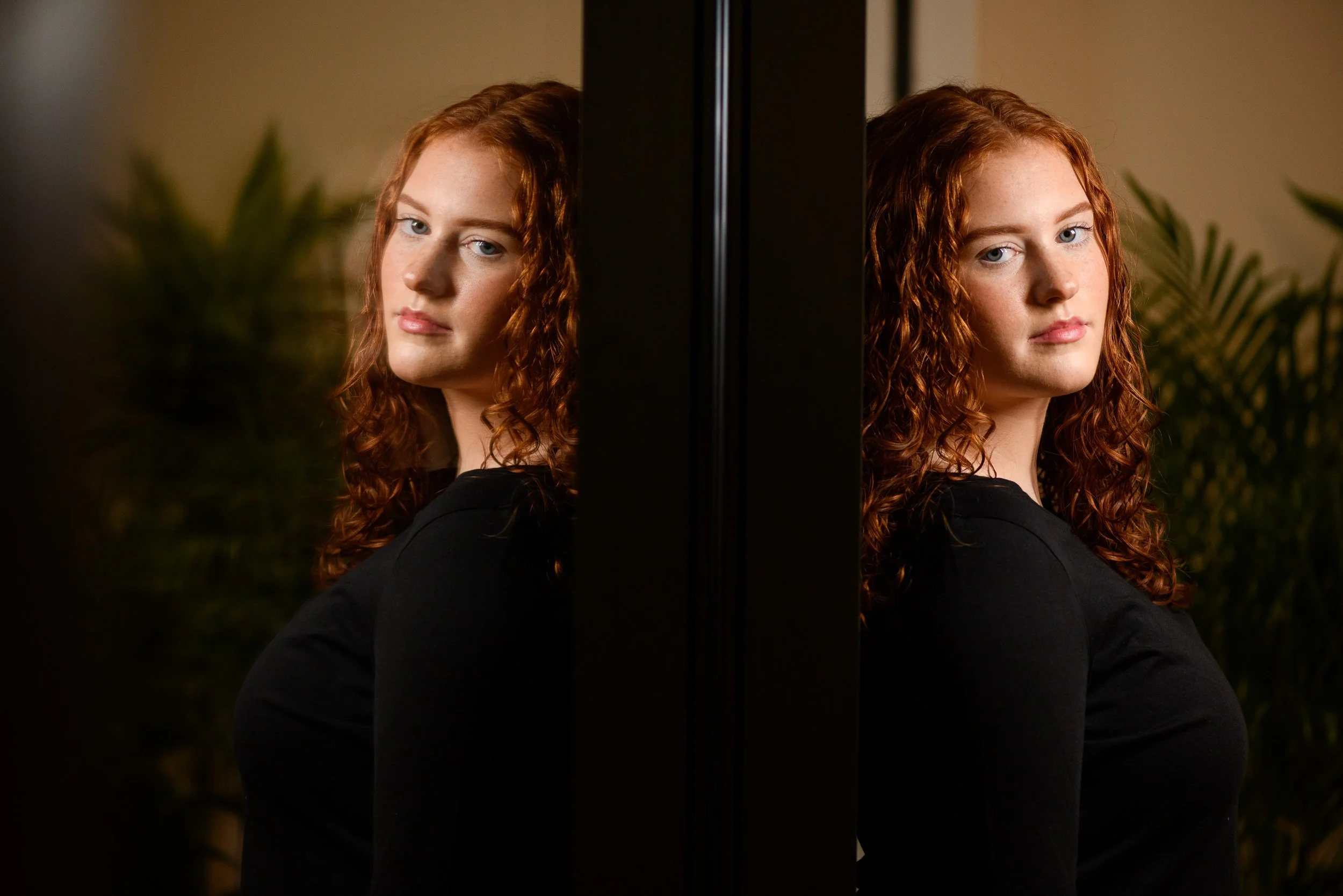 Portrait of a young woman with curly red hair reflected in a mirror with greenery

