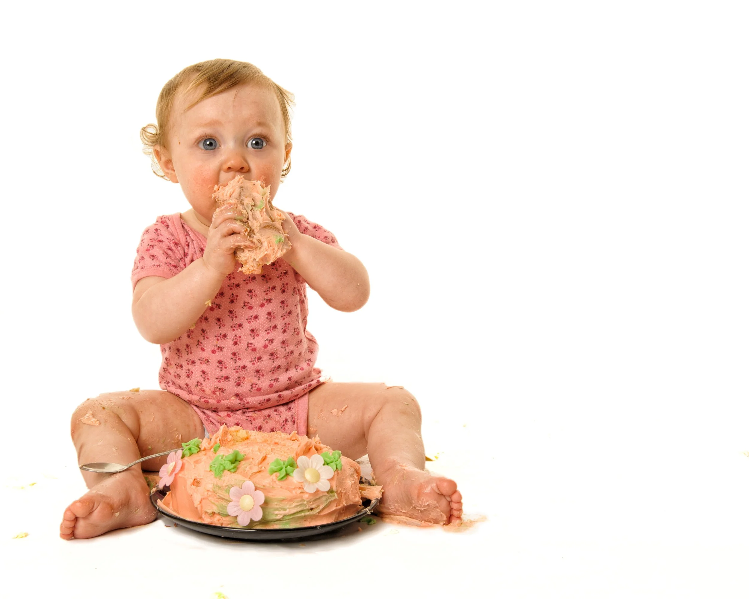 A young child with blue eyes and curly blonde hair, wearing a pink floral shirt, sitting on the floor with a large pink cake decorated with flowers, eating the cake with both hands.