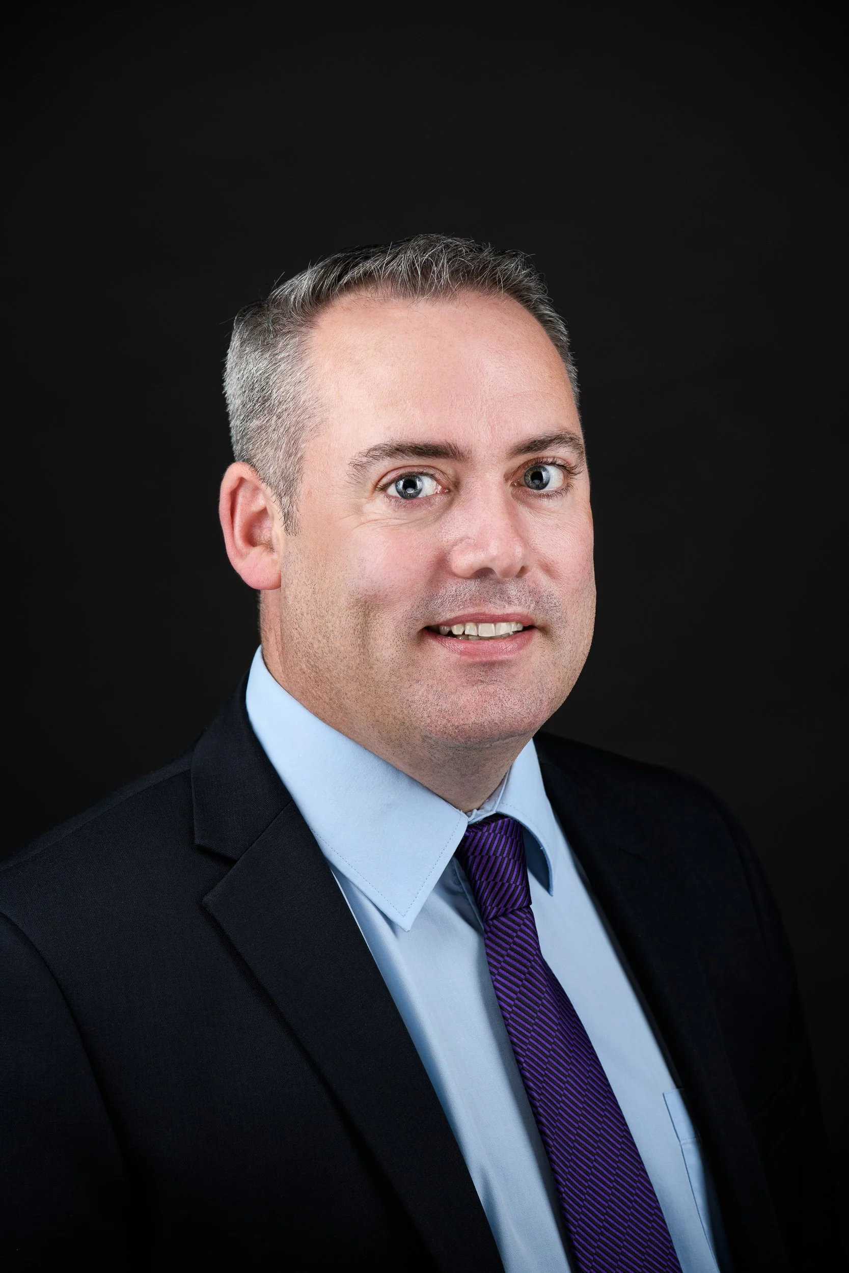 Headshot of a man in a business suit, posed confidently against a dark background.
