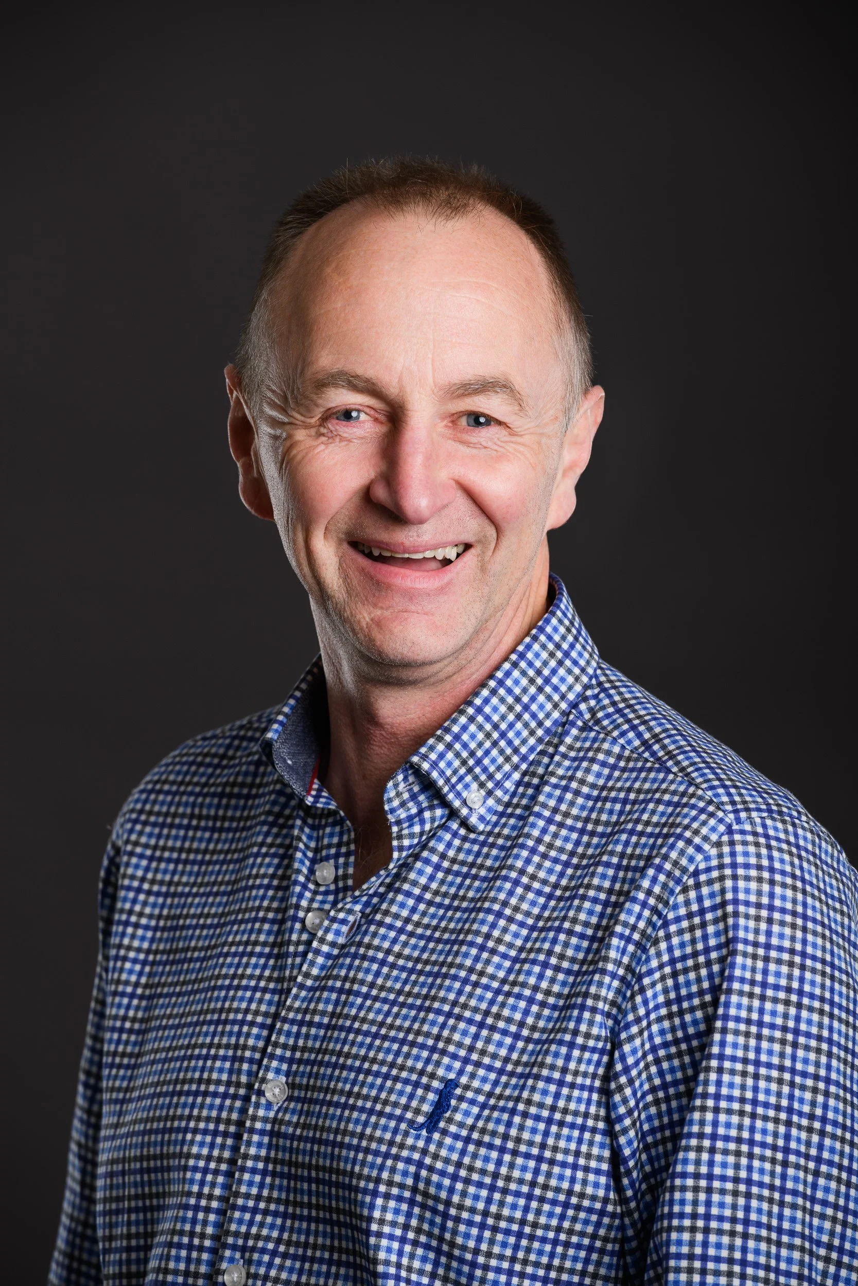 Portrait of a smiling middle-aged man wearing a blue and white checkered shirt against a dark background.
