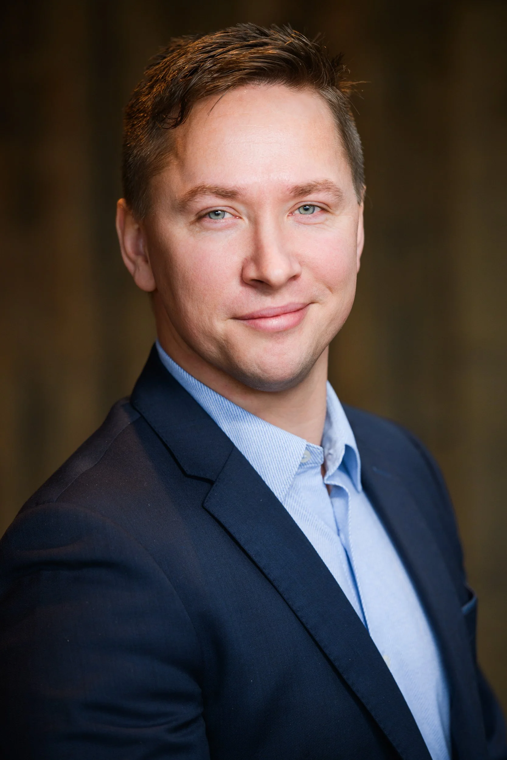 Close-up headshot of a young man in a navy suit and light blue shirt, smiling subtly toward the camera.
