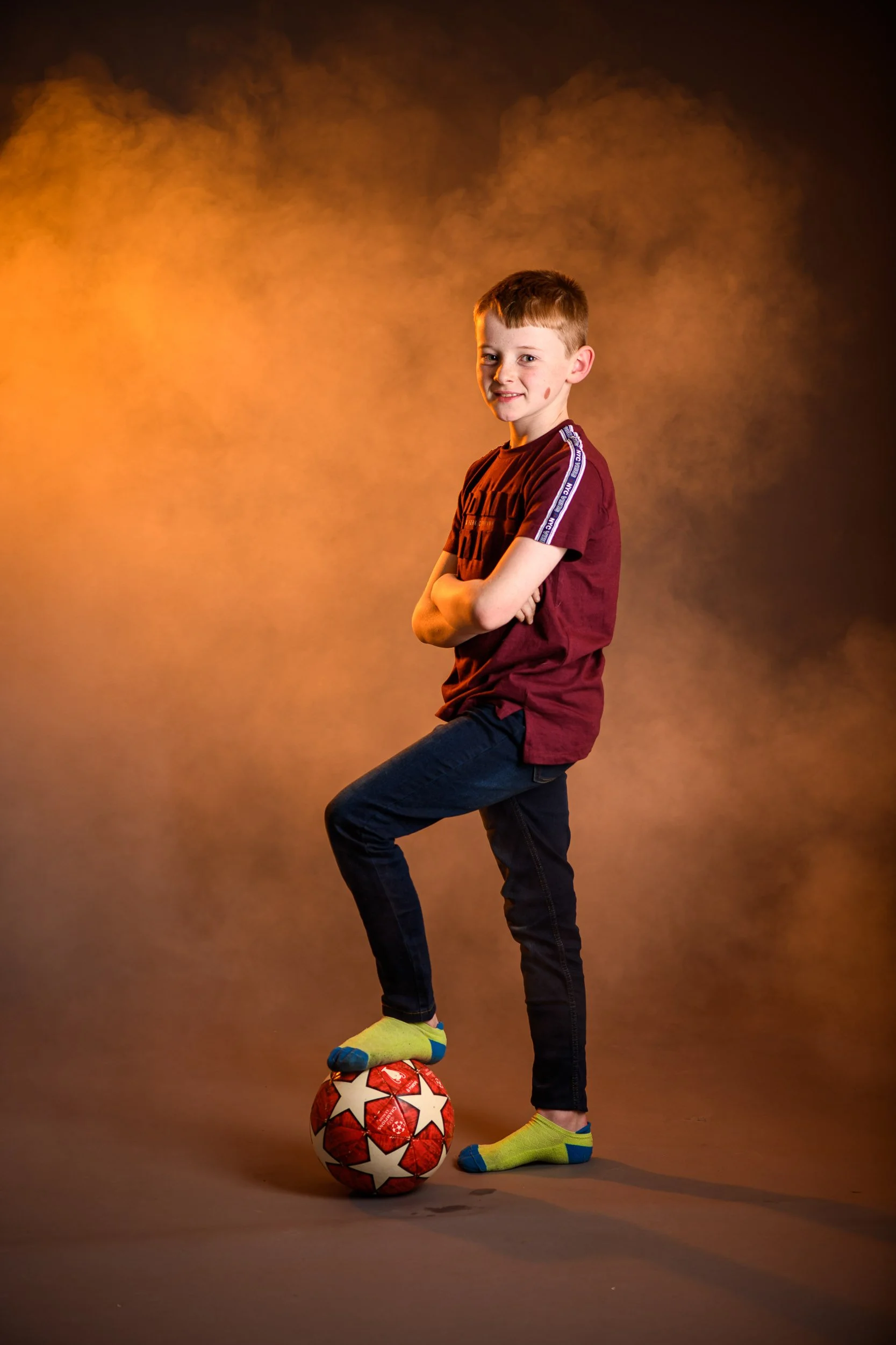 A young boy with a football, standing on one foot while the other foot rests on the ball, against a warm, smoky background, wearing yellow socks with blue tips, dark jeans, and a maroon T-shirt.