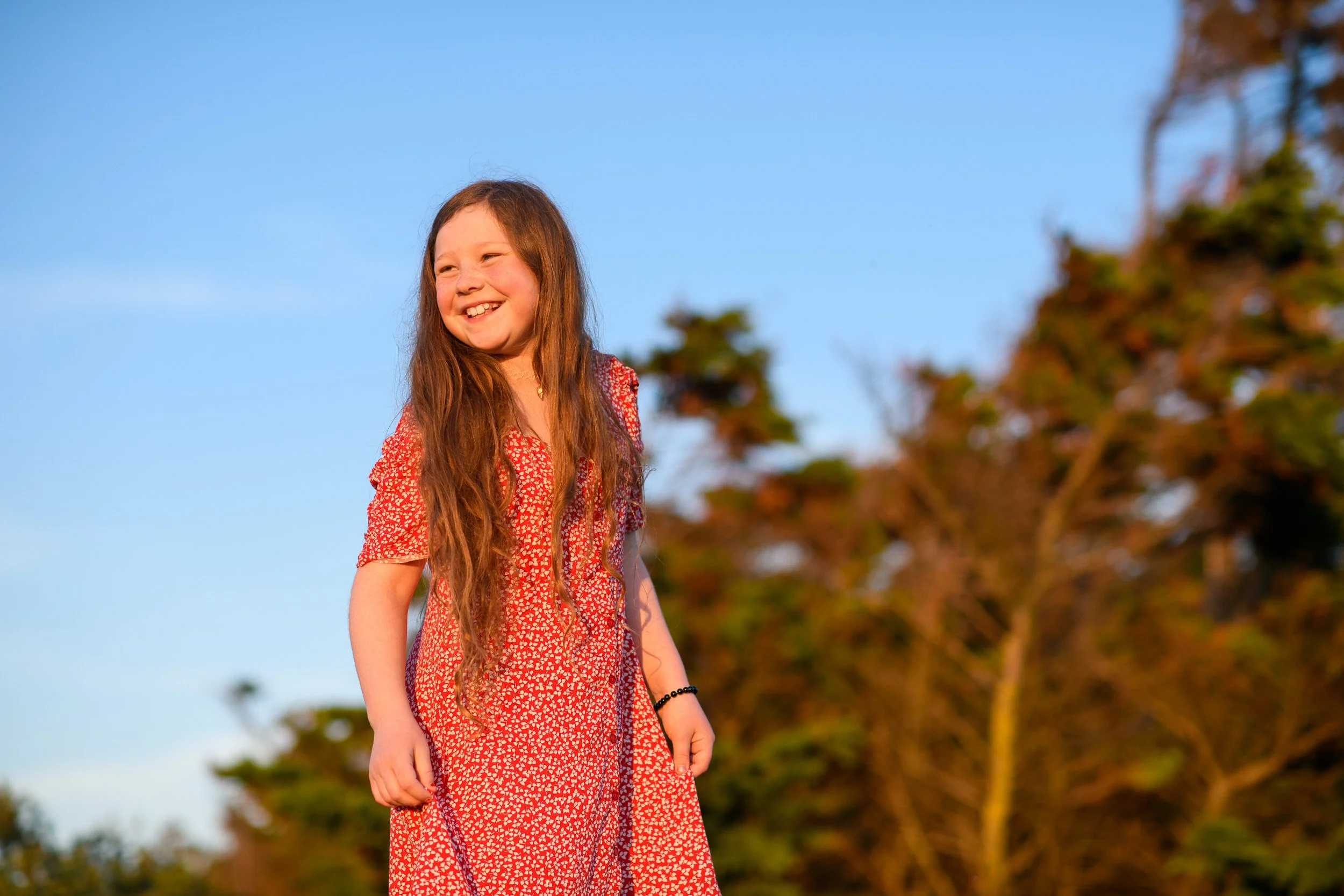 A young girl smiling outdoors during sunset with trees softly blurred in the background.