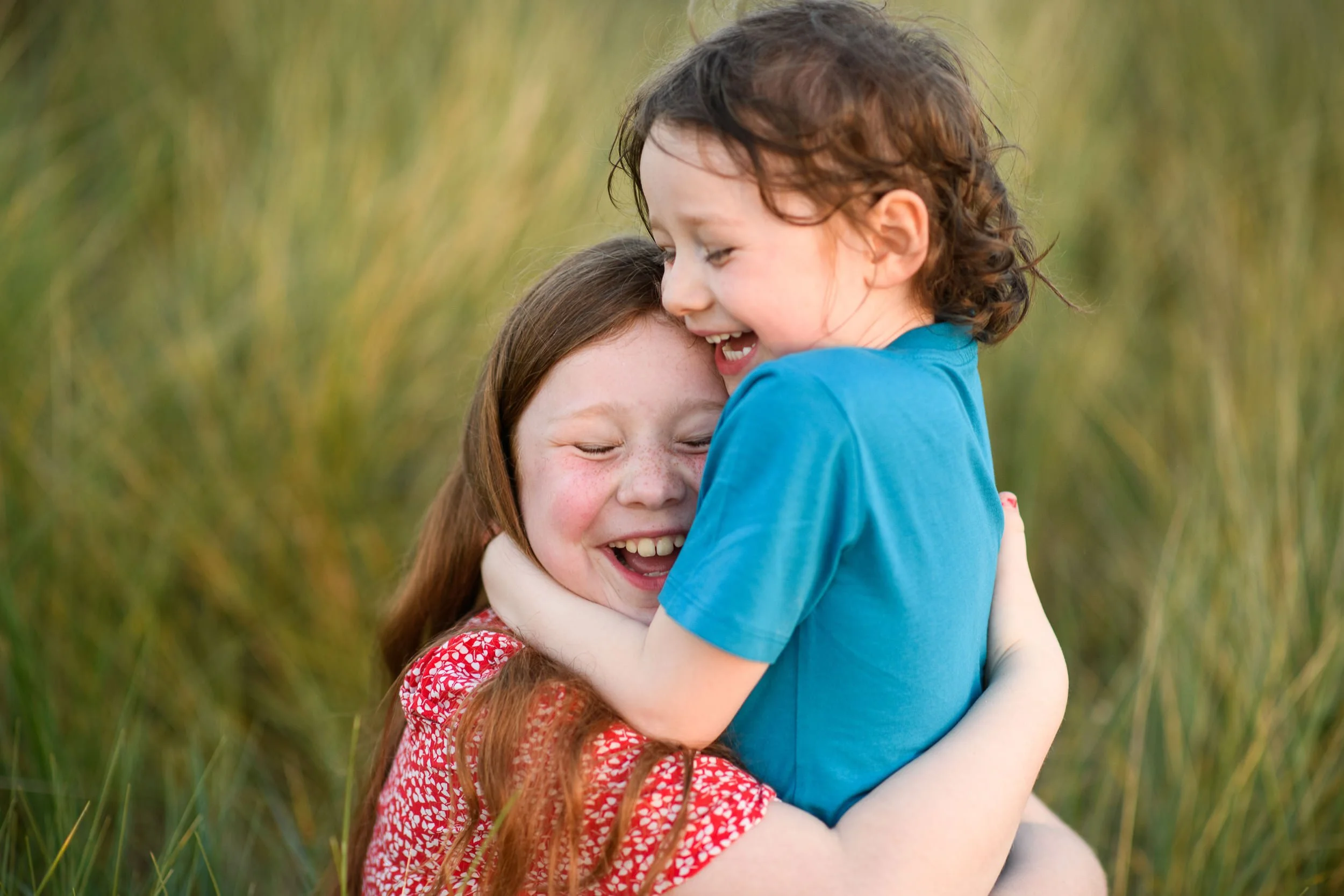 Two children hugging and laughing outdoors with blurred green grass background.