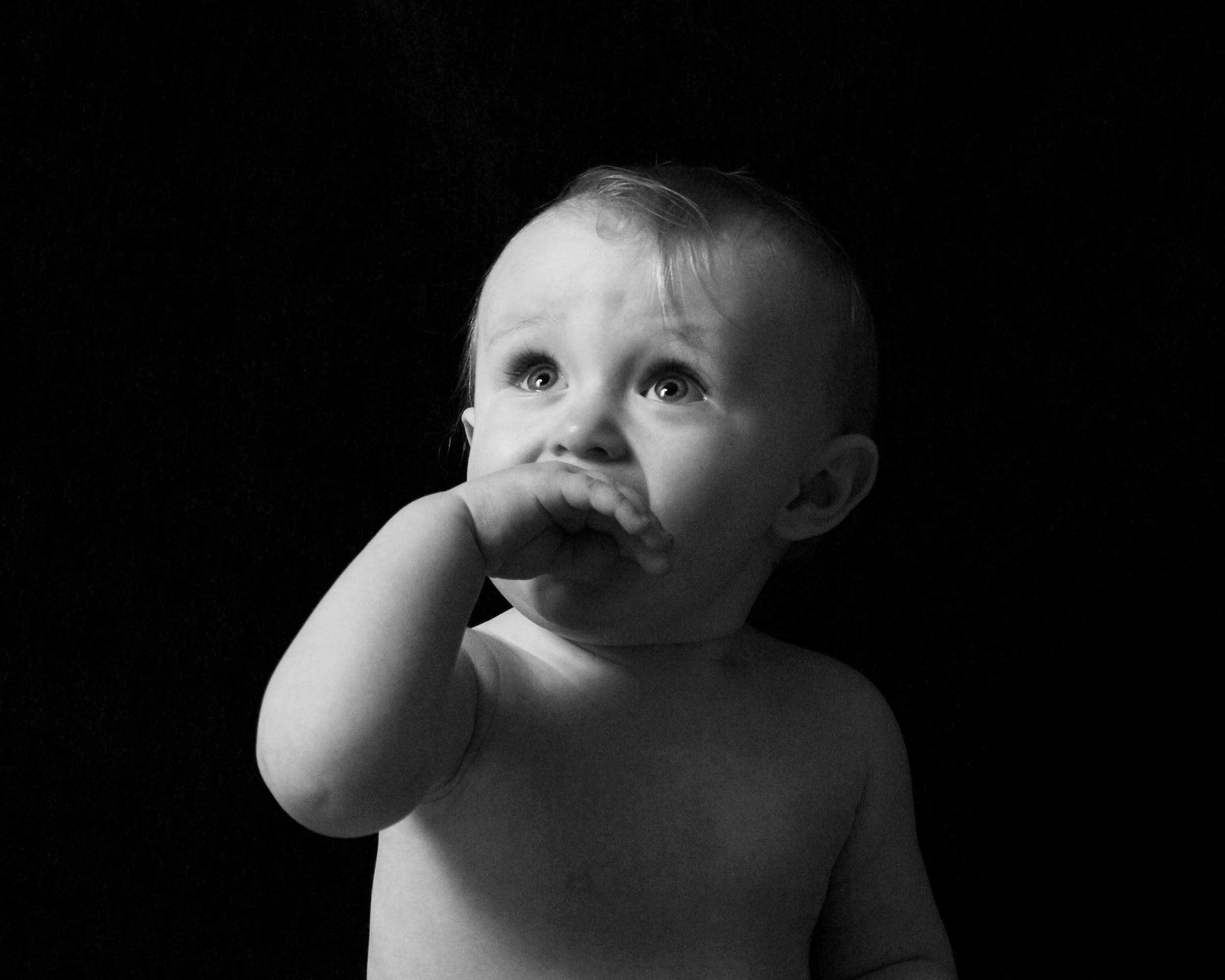 Black and white portrait of a young child looking upward with their hand near their mouth.

