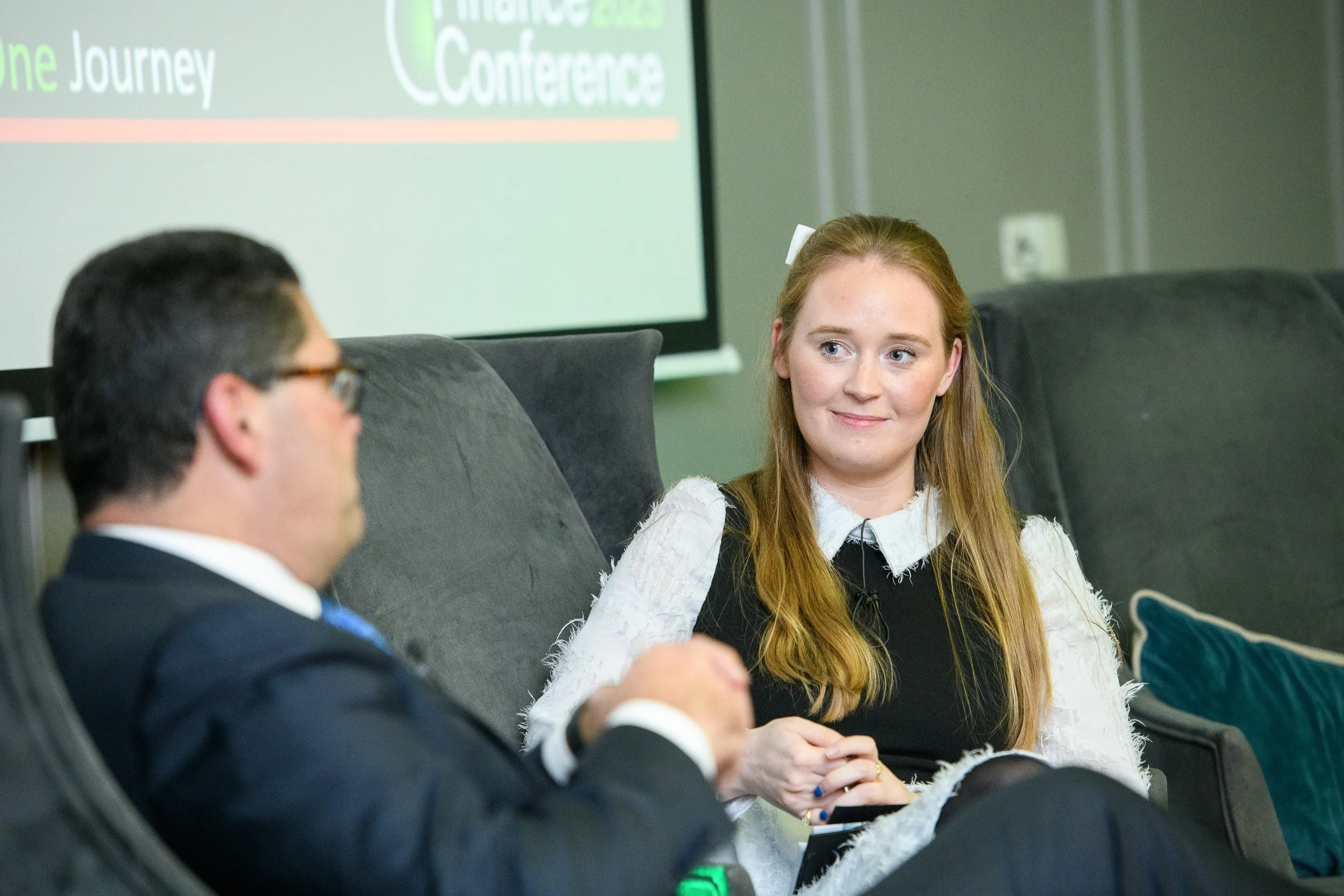A woman seated on a sofa listening attentively to a speaker during a conference setting.
