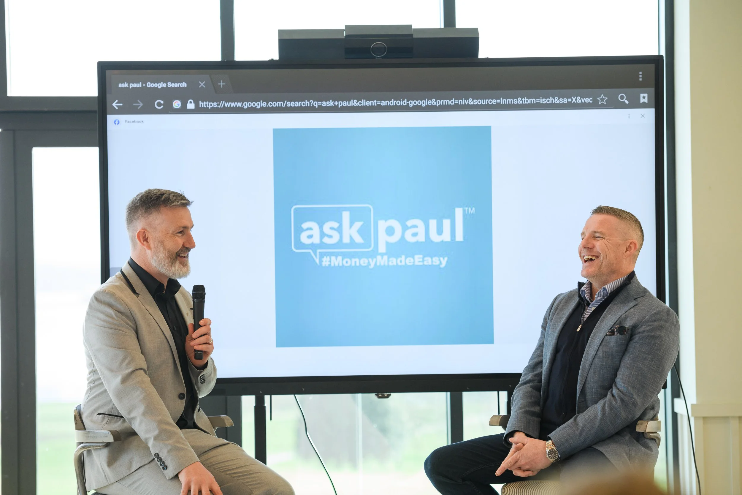 Two men sitting and laughing during a presentation, with a large screen behind them displaying the logo 'ask paul' and the hashtag '#MoneyMadeEasy'.
