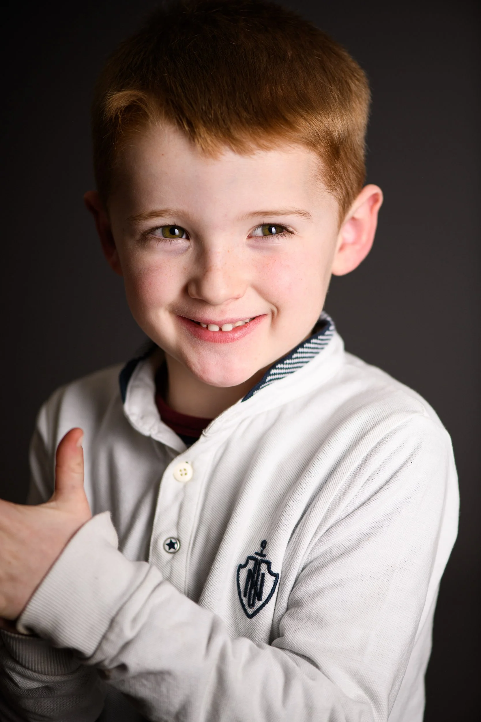 Young boy with red hair and freckles, smiling and giving a thumbs-up, wearing a white shirt with a crest on the chest, against a dark background.