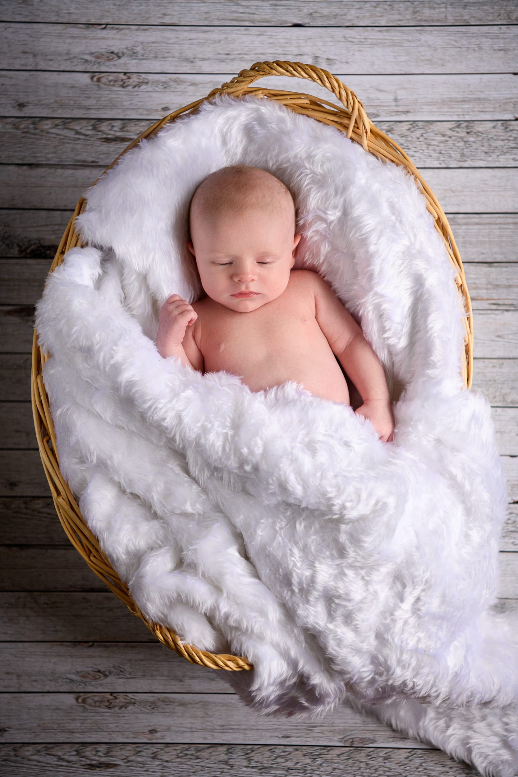 A baby sleeping in a wicker basket lined with white fluffy blanket on a wooden floor.