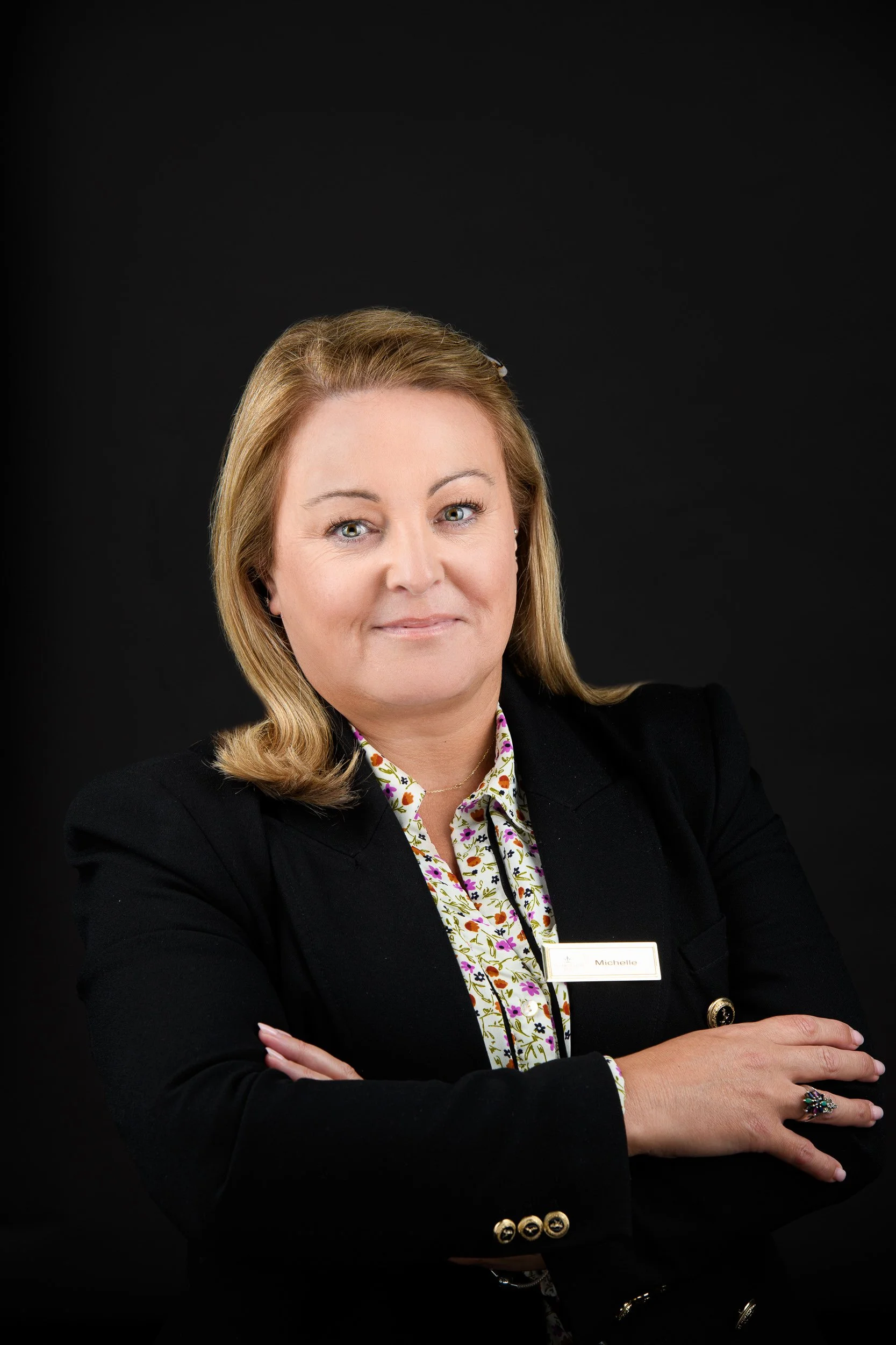 Professional headshot of a woman with blonde hair wearing a black blazer and floral shirt, standing with arms crossed against a dark background.
