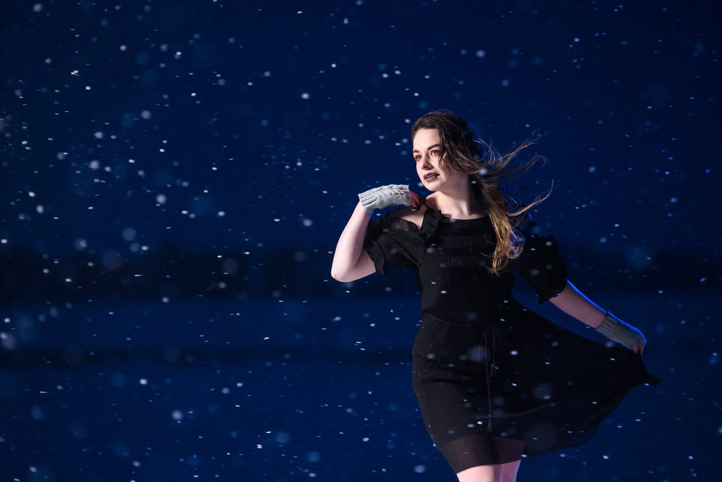 Cinematic portrait of a woman standing in falling snow at night wearing a black dress and gloves

