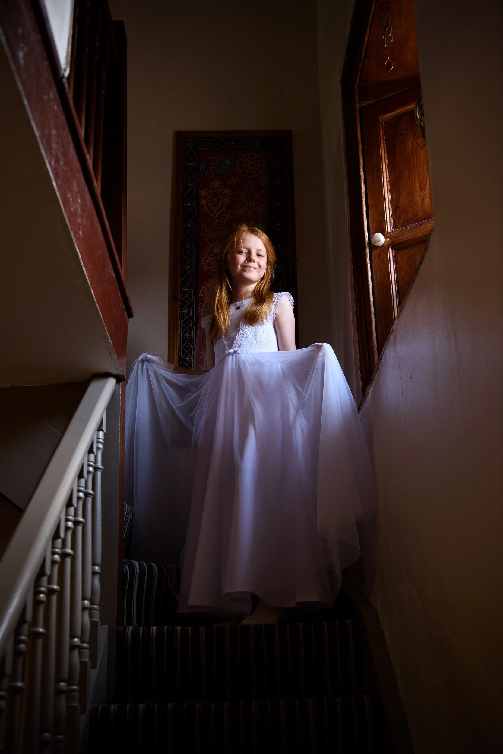 A young girl with red hair wearing a white dress standing at the top of a staircase.