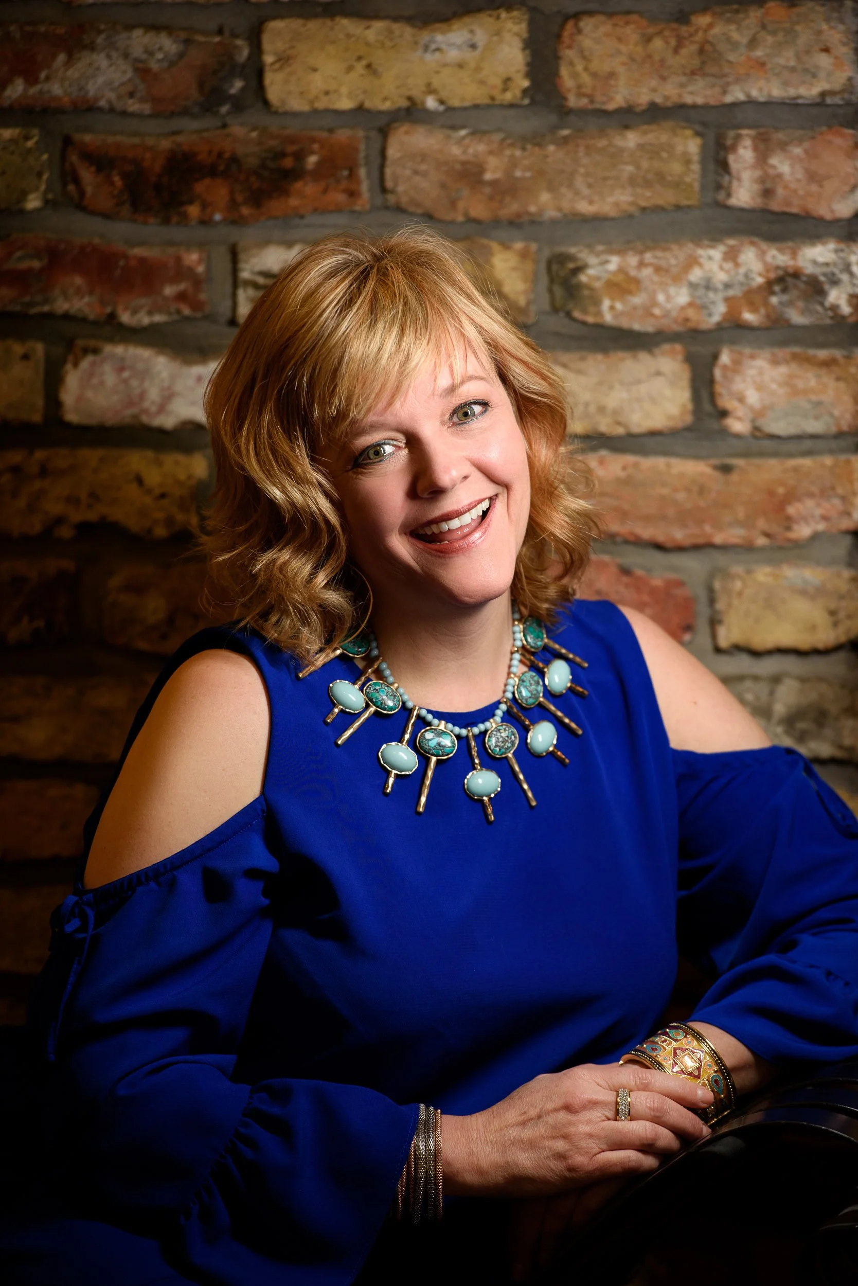 Portrait of a woman with curly red hair wearing a blue top and statement necklace, seated against a textured brick wall.
