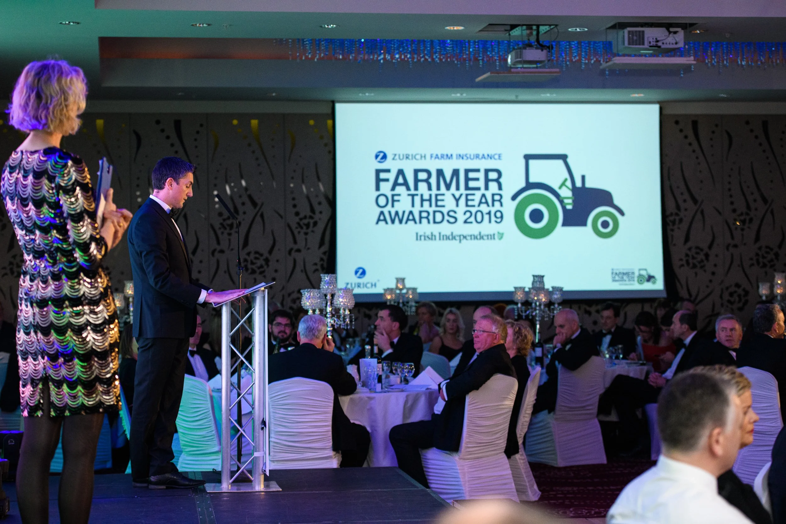 An award ceremony with a large screen displaying 'Farmer of the Year Awards 2019' by Zurich Farm Insurance. Attendees seated at tables, with two presenters on stage, one woman in a colorful dress and one man in a black suit, speaking at a podium.
