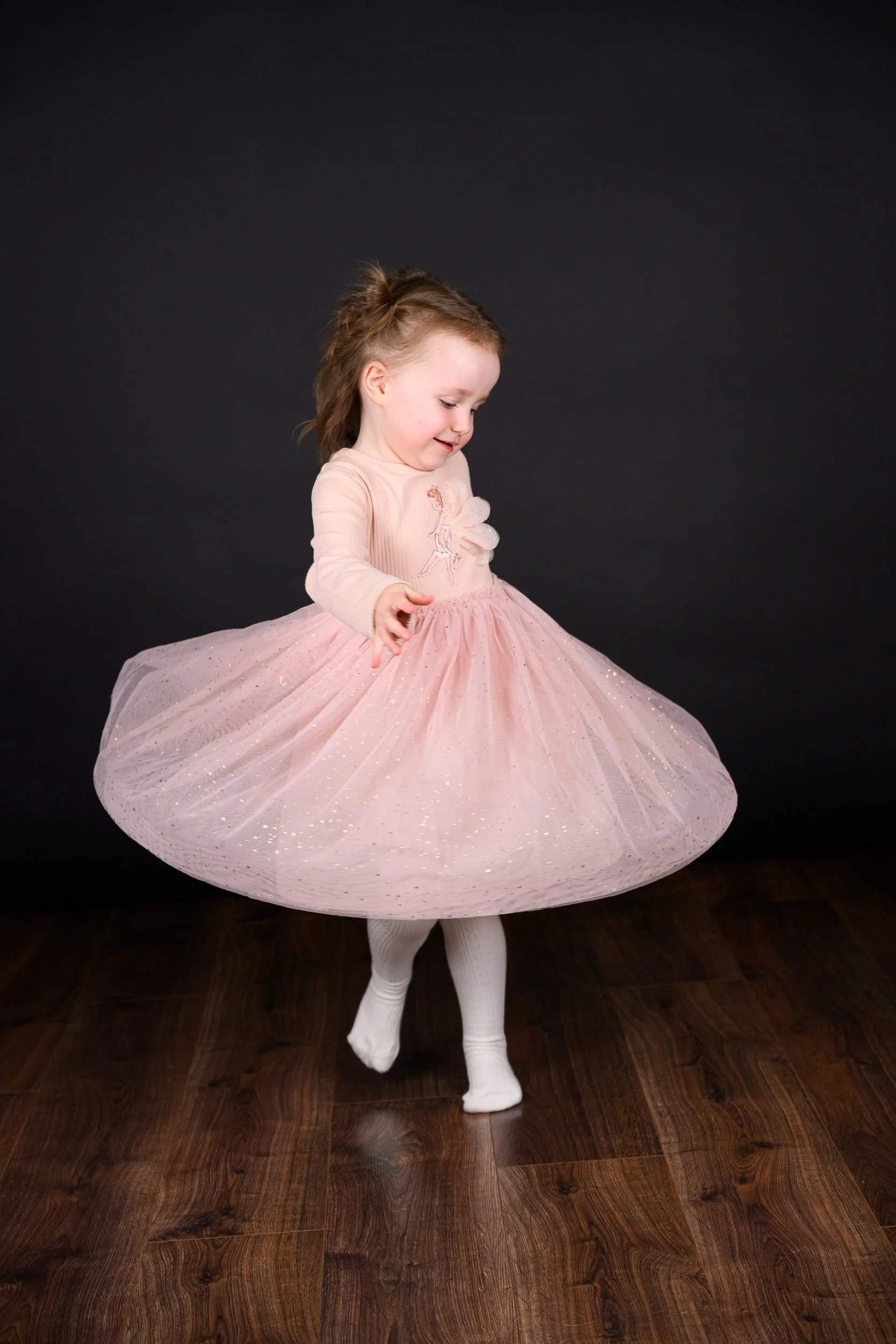 A young girl in a pink dress smiling and looking at the camera against a black background.
