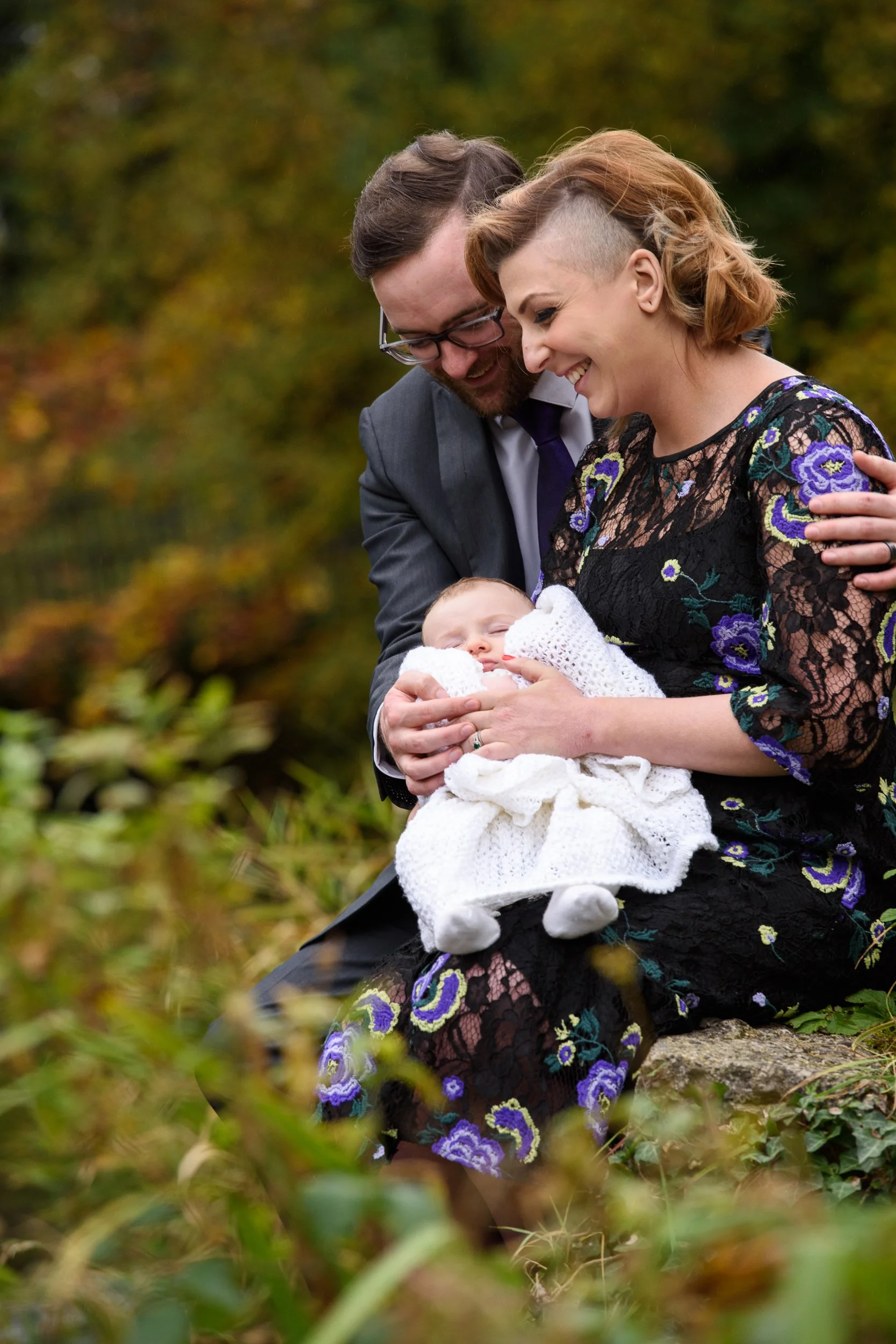 A couple holding their sleeping baby outdoors with autumn foliage in the background.
