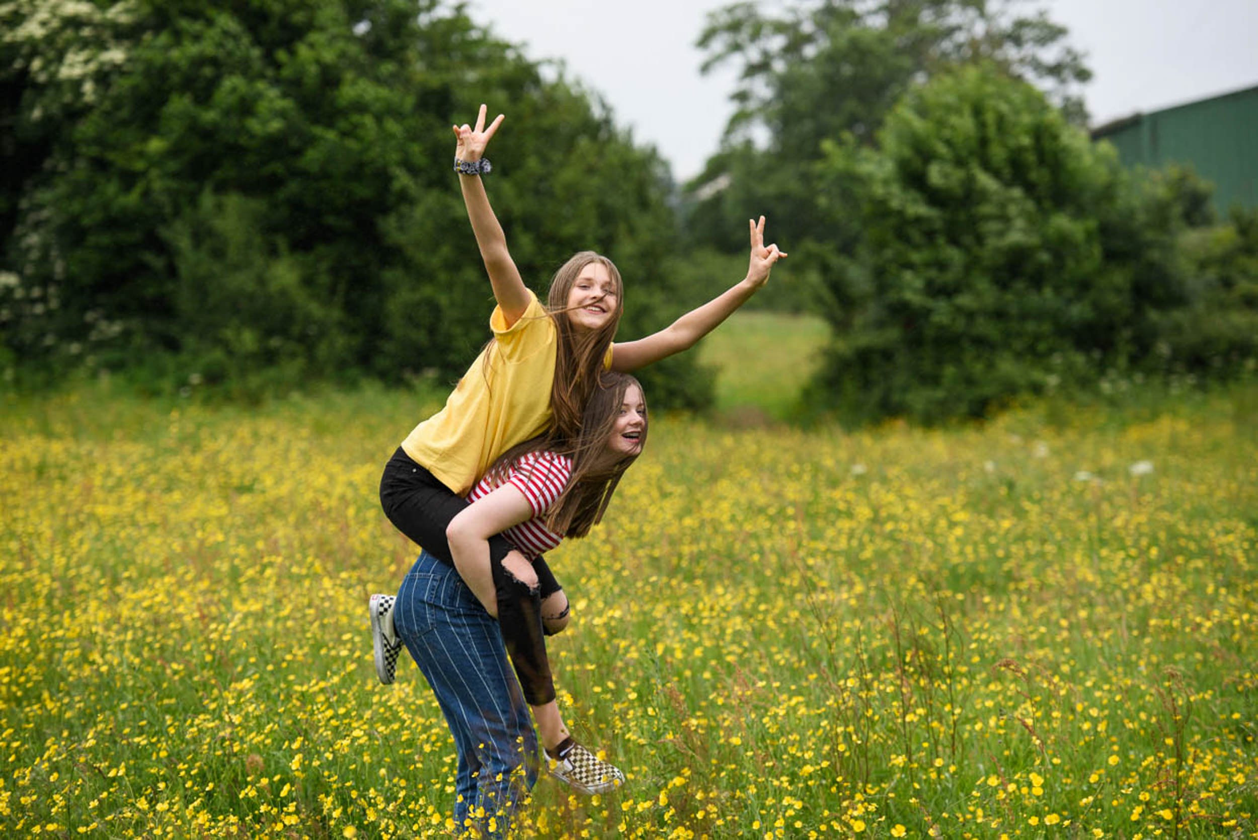 Two young women smiling and playing in a field of yellow flowers, one giving the other a piggyback ride. They are outdoors with trees and a building in the background.