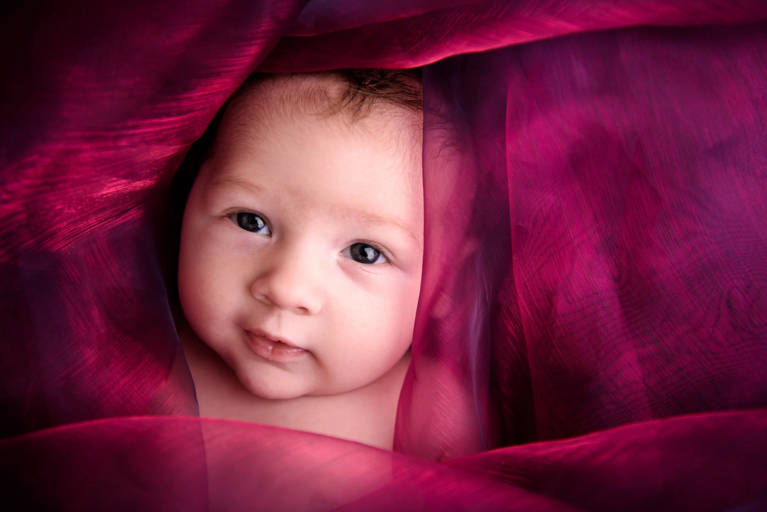 Close-up of a baby with blue eyes and light skin peeking through deep pink and purple translucent fabric, smiling gently.