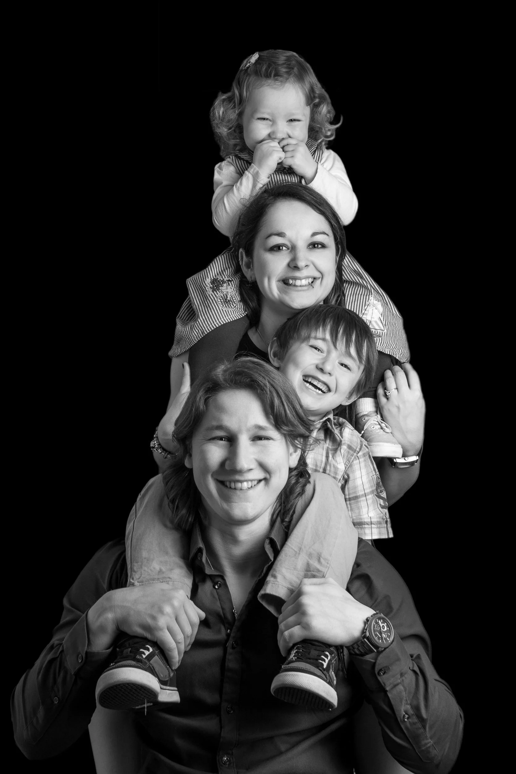 Black and white portrait of a family of four arranged in a playful stacked pose against a dark background.
