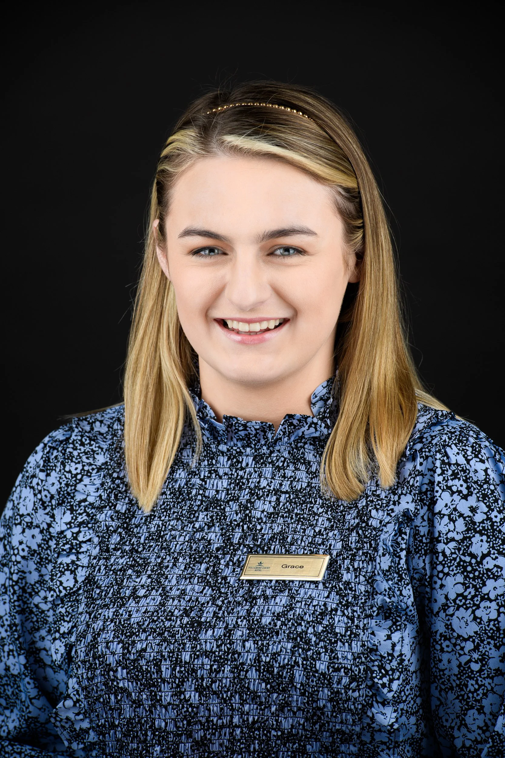 Close-up portrait of a smiling woman with blonde hair wearing a blue floral uniform and name badge, set against a black background.