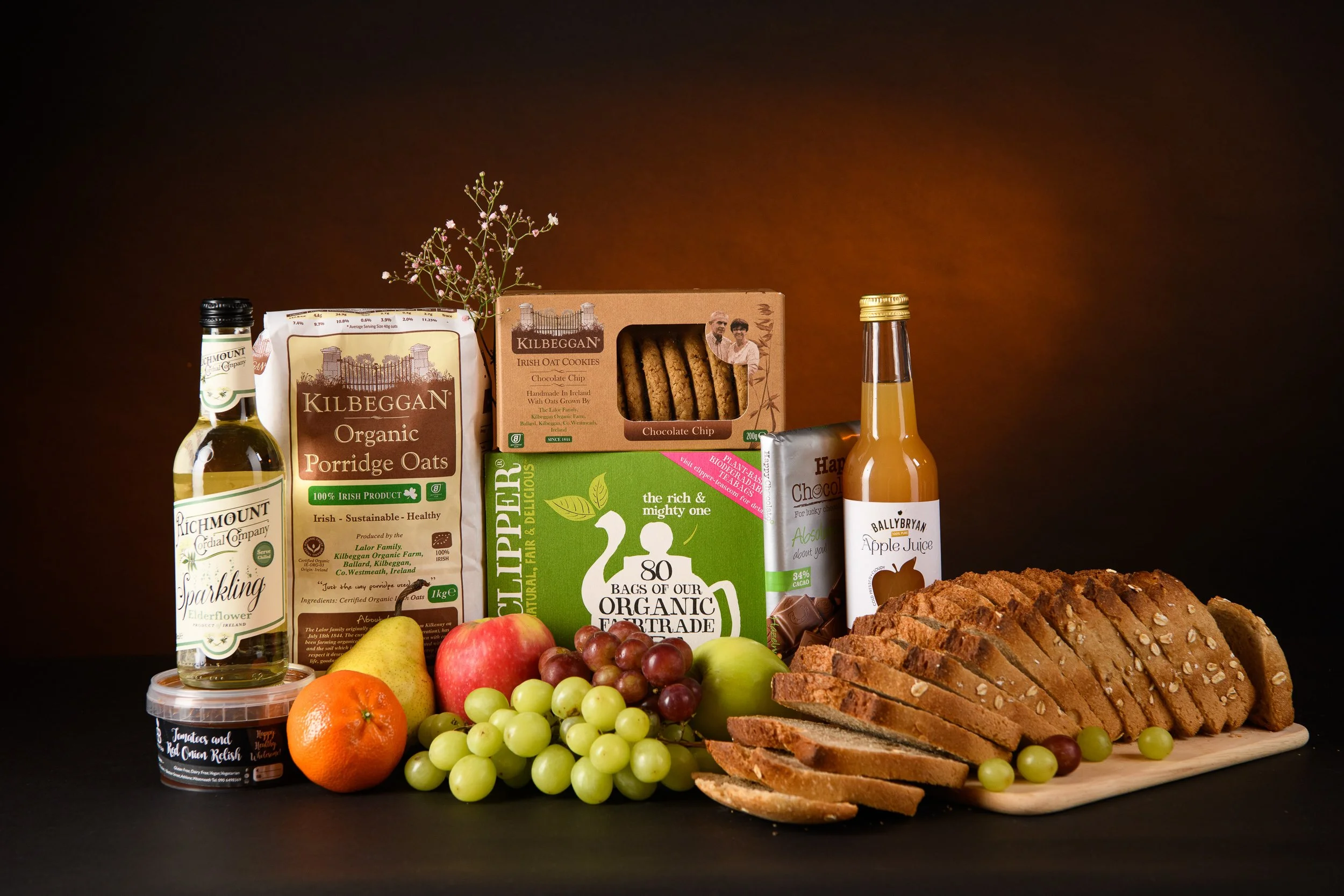 A variety of food items including sliced bread on a cutting board, grapes, an apple, an orange, a pear, a box of organic porridge oats, a package of Irish oat cookies, a bottle of apple juice, a bottle of elderflower syrup, a jar of tomato and red on