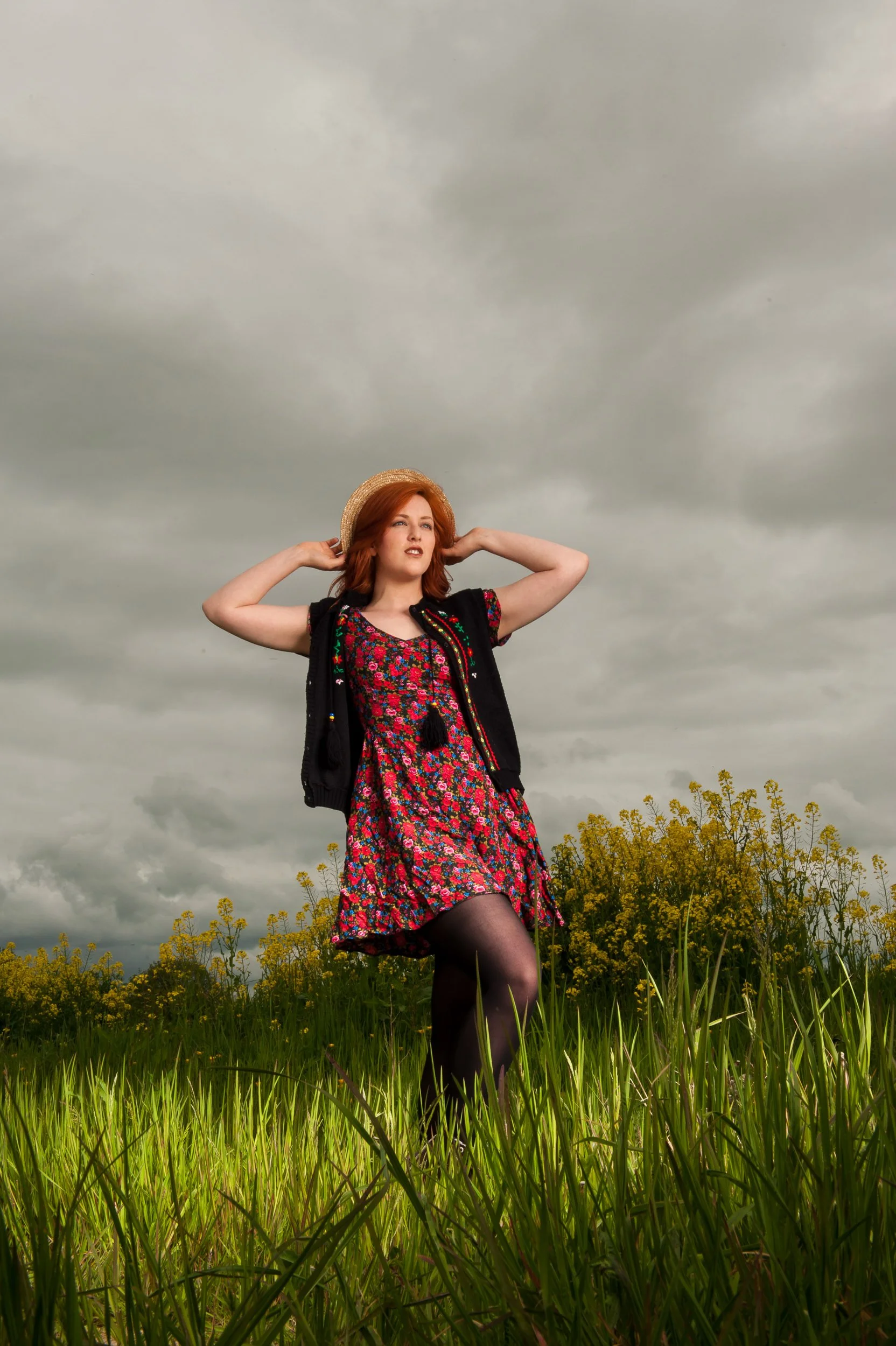 Outdoor portrait of a woman with red hair in a field wearing a floral dress and holding a straw hat
