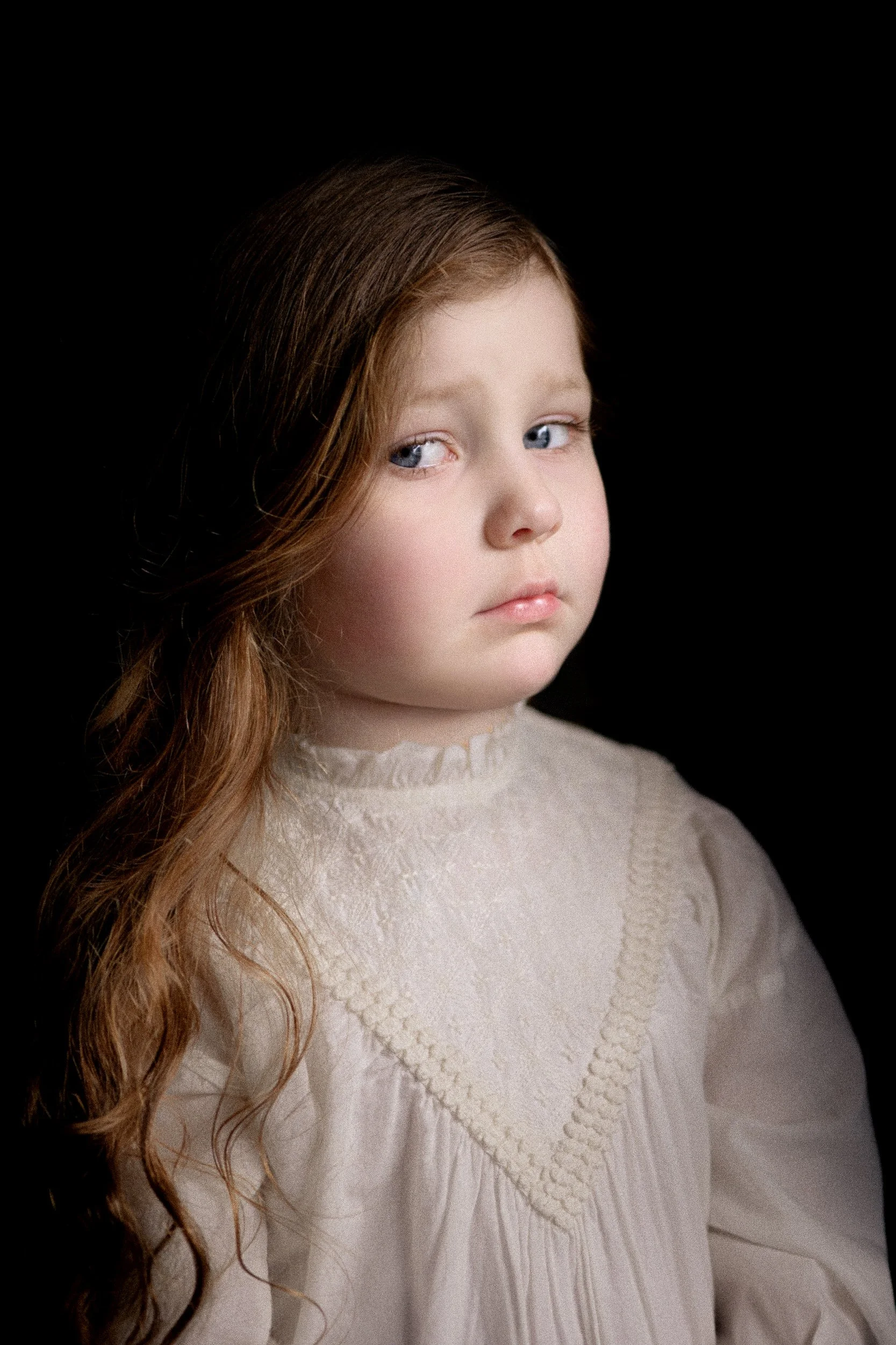 Ken Byrne Portrait Photography - A young girl with long red hair and blue eyes wearing a lace dress against a black background.
