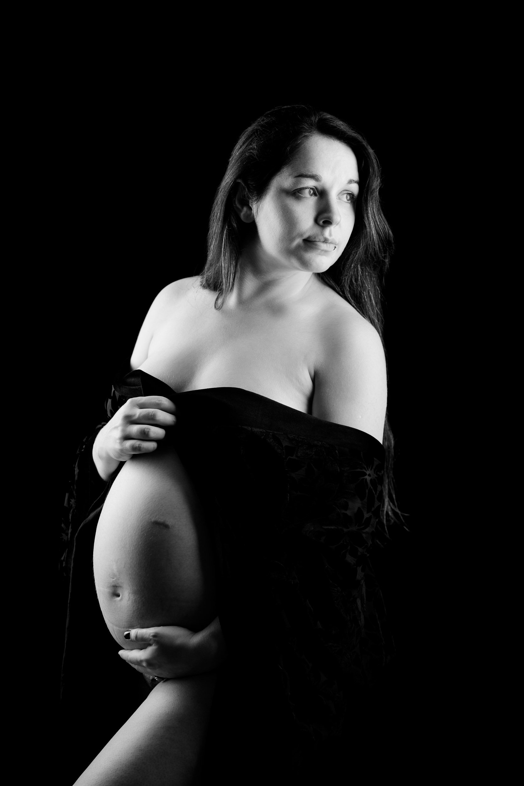 Black and white portrait of a pregnant woman holding her belly in profile against a dark background.
