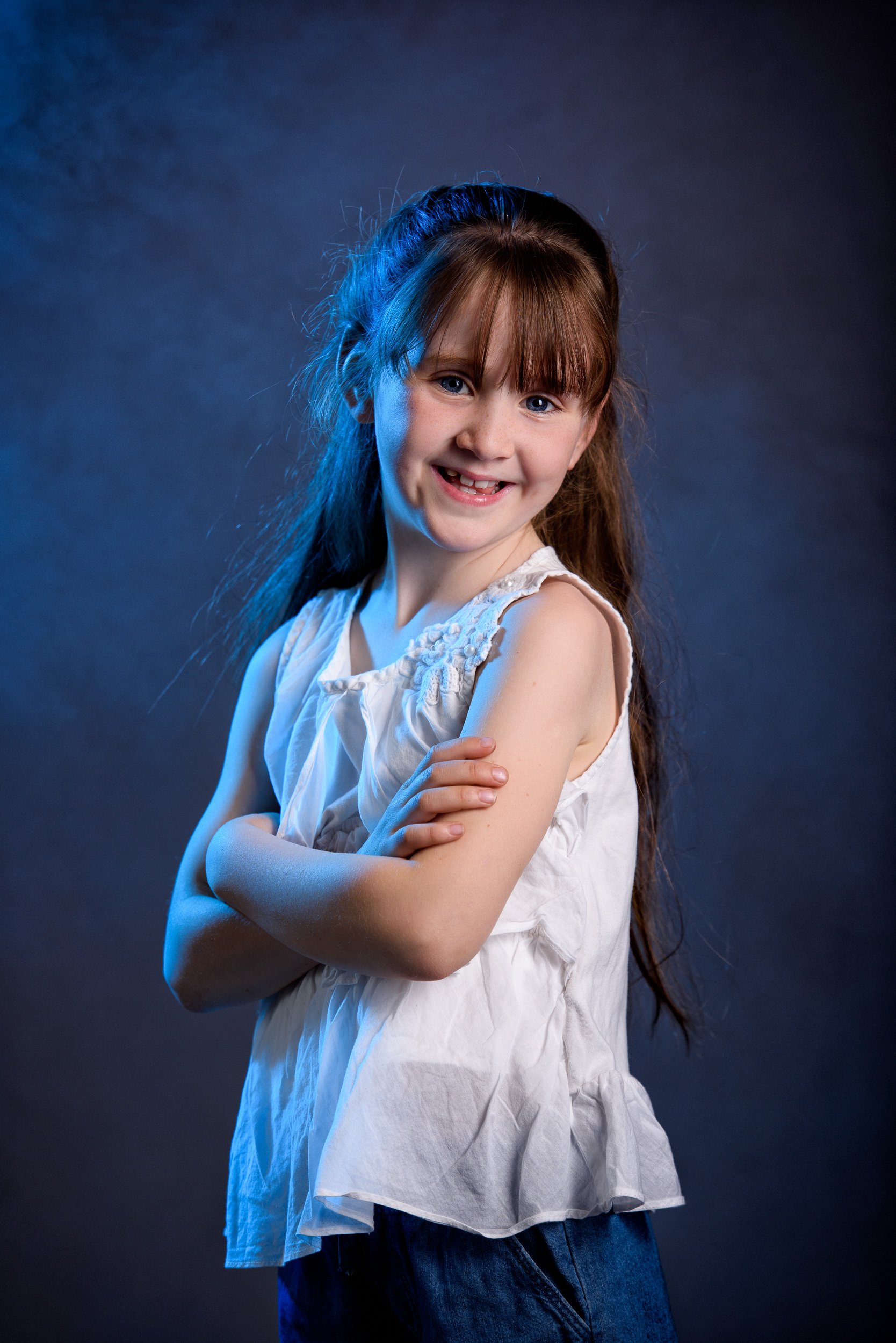 A young girl with long brown hair, smiling, wearing a white sleeveless top and blue jeans, standing against a dark background with blue lighting.