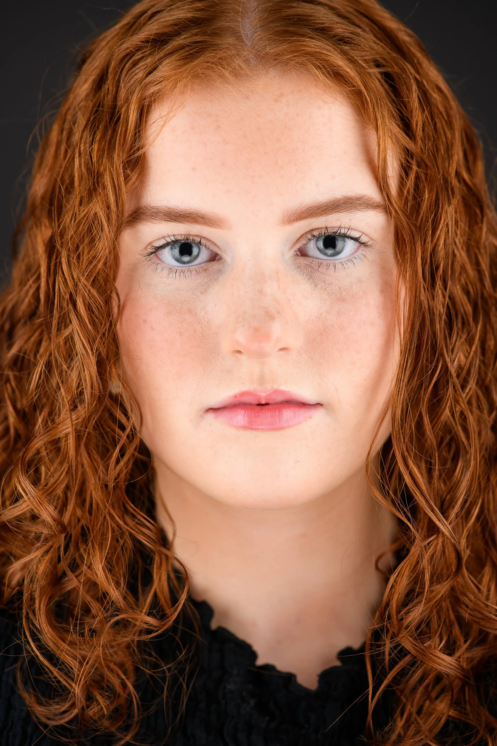Close-up portrait of a young woman with curly red hair and blue eyes against a dark background.