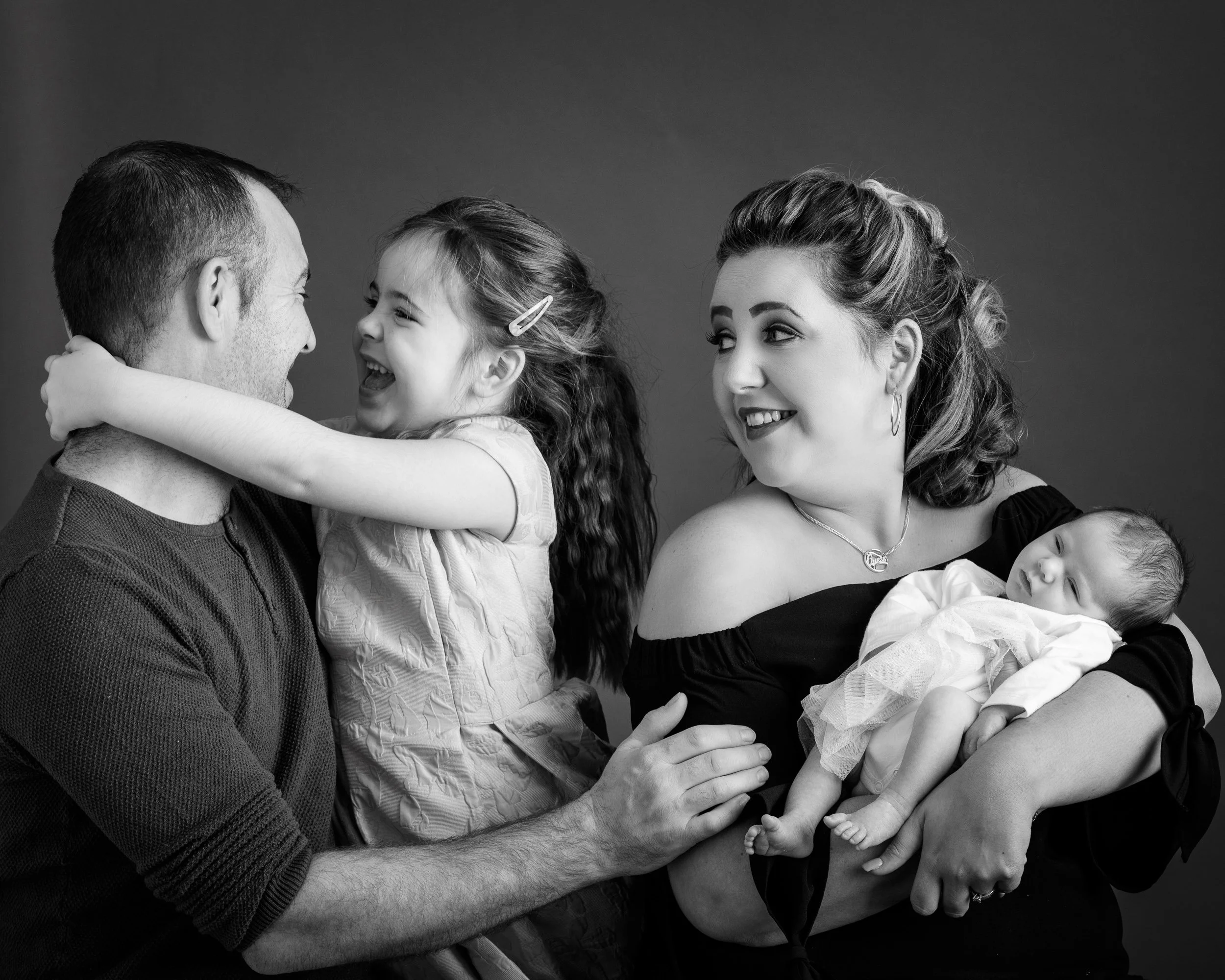 Black and white family portrait of parents with a young girl and sleeping baby, sharing a warm moment together.
