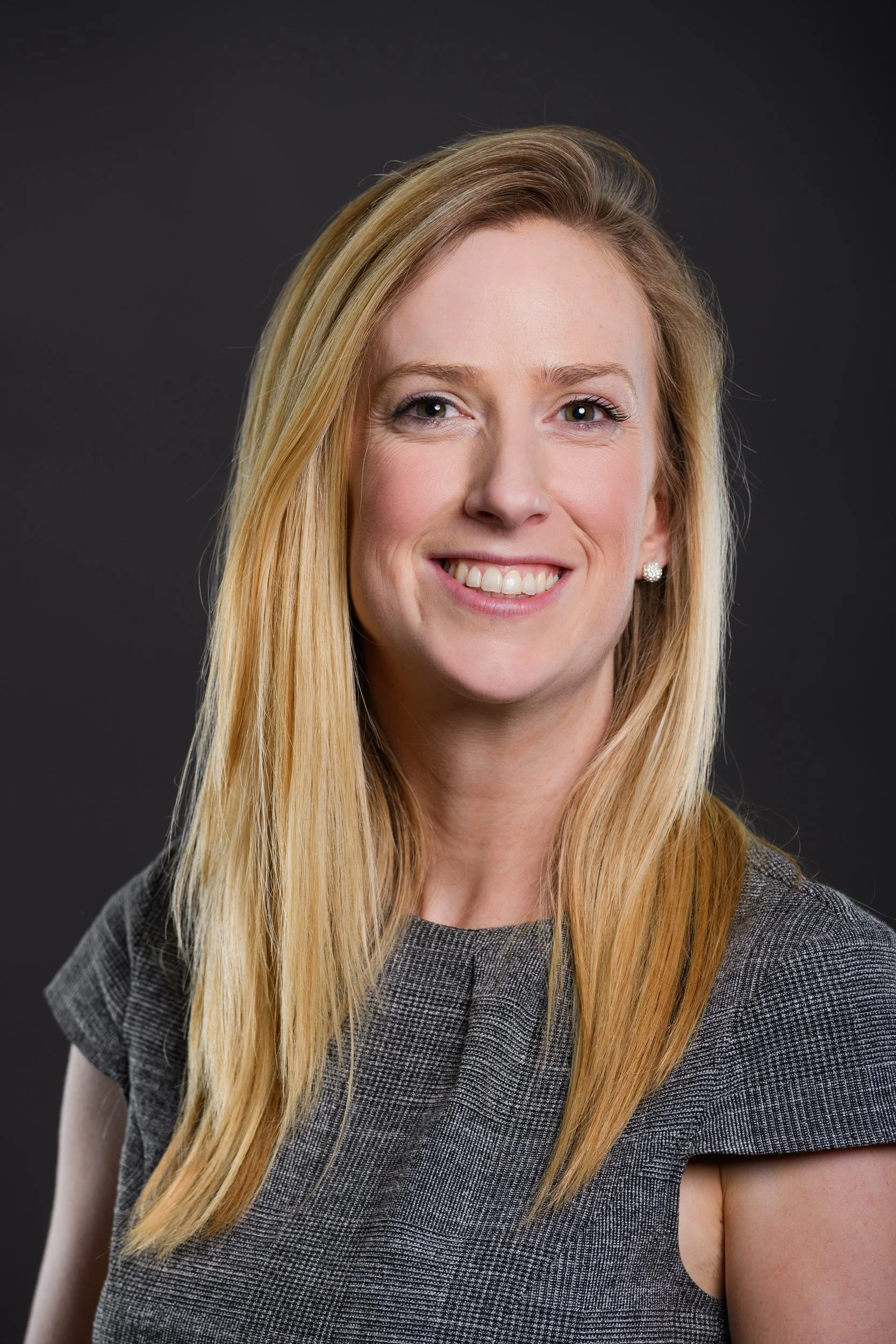 Portrait of a smiling woman with long blonde hair wearing a grey checkered top, posed against a dark grey background.
