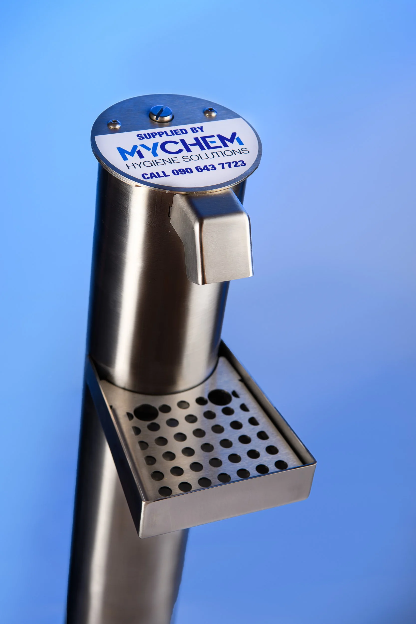 Wall-mounted stainless steel hand sanitiser dispenser with branded label against a light blue background.
