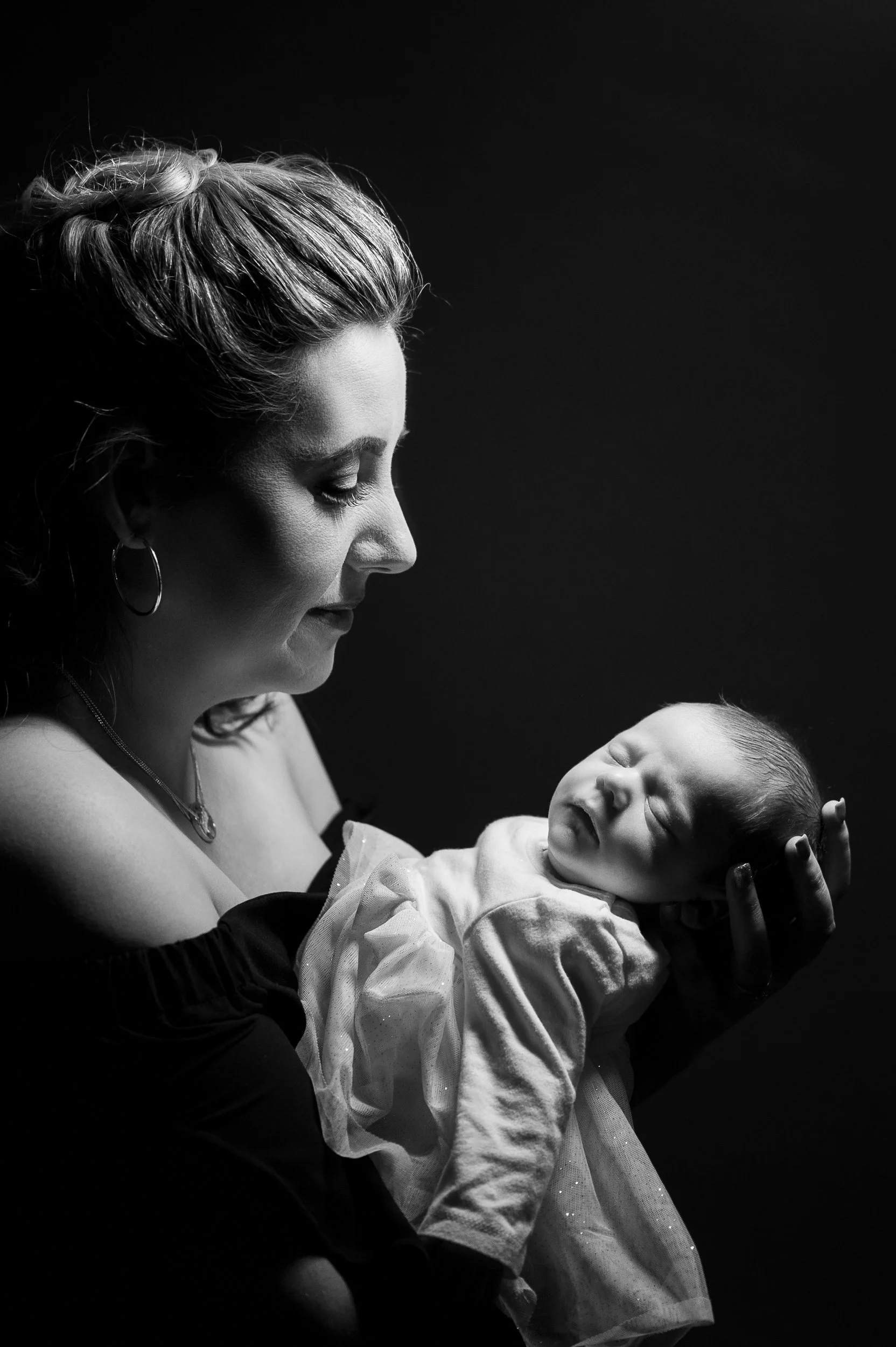 A black and white portrait of a woman holding a newborn baby close to her chest, both in profile view, with a dark background.
