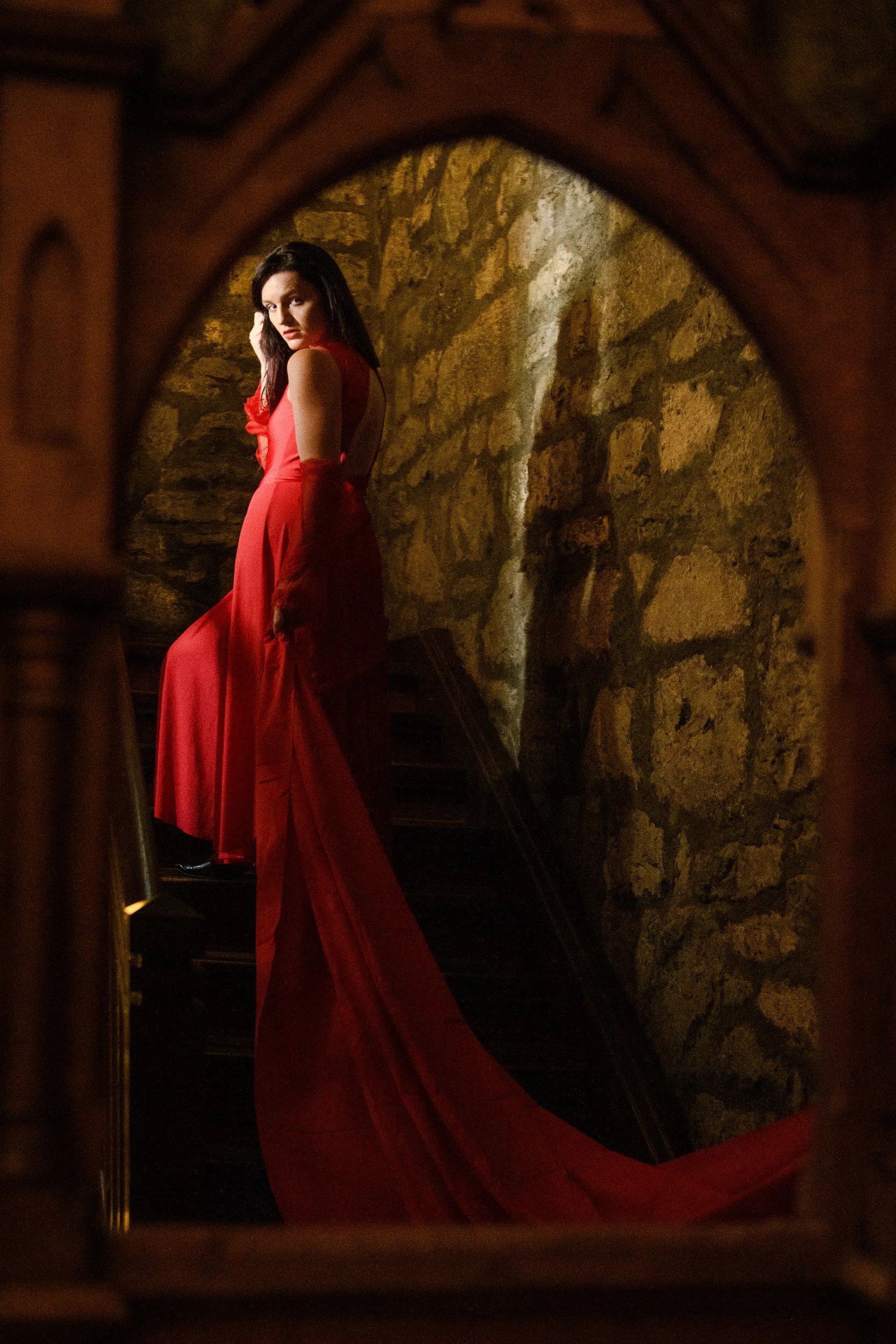 A woman in a red evening gown with a long train standing on a staircase, viewed through a wooden frame, against a stone wall background.