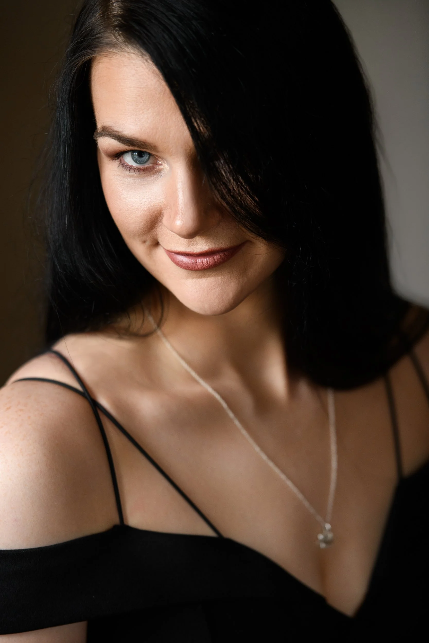 A woman with long dark hair and blue eyes, wearing a black dress and silver necklace, posing with a soft smile against a neutral indoor background.