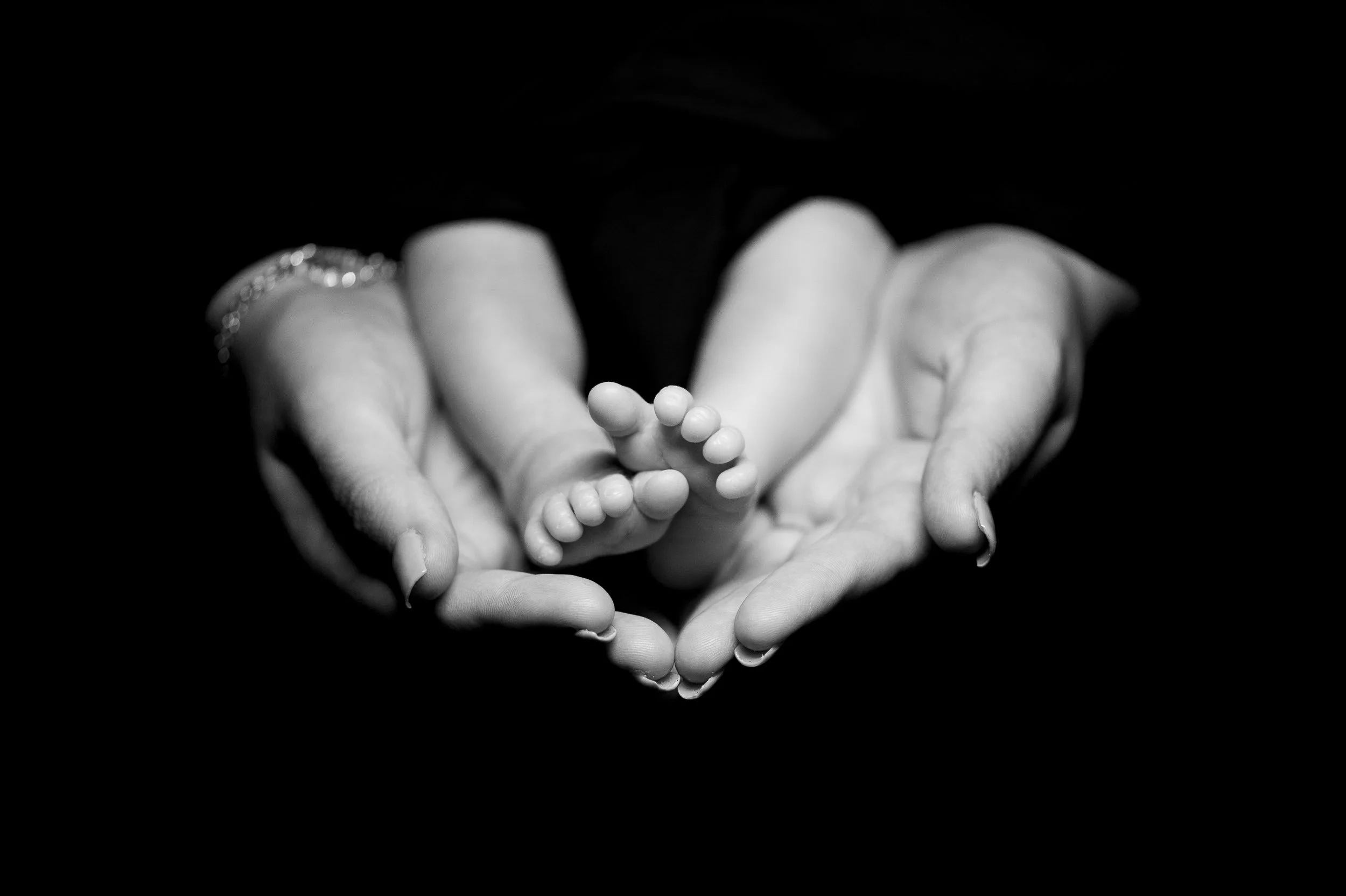 Black and white photograph of adult hands gently holding a tiny baby's feet, forming a heart shape.
