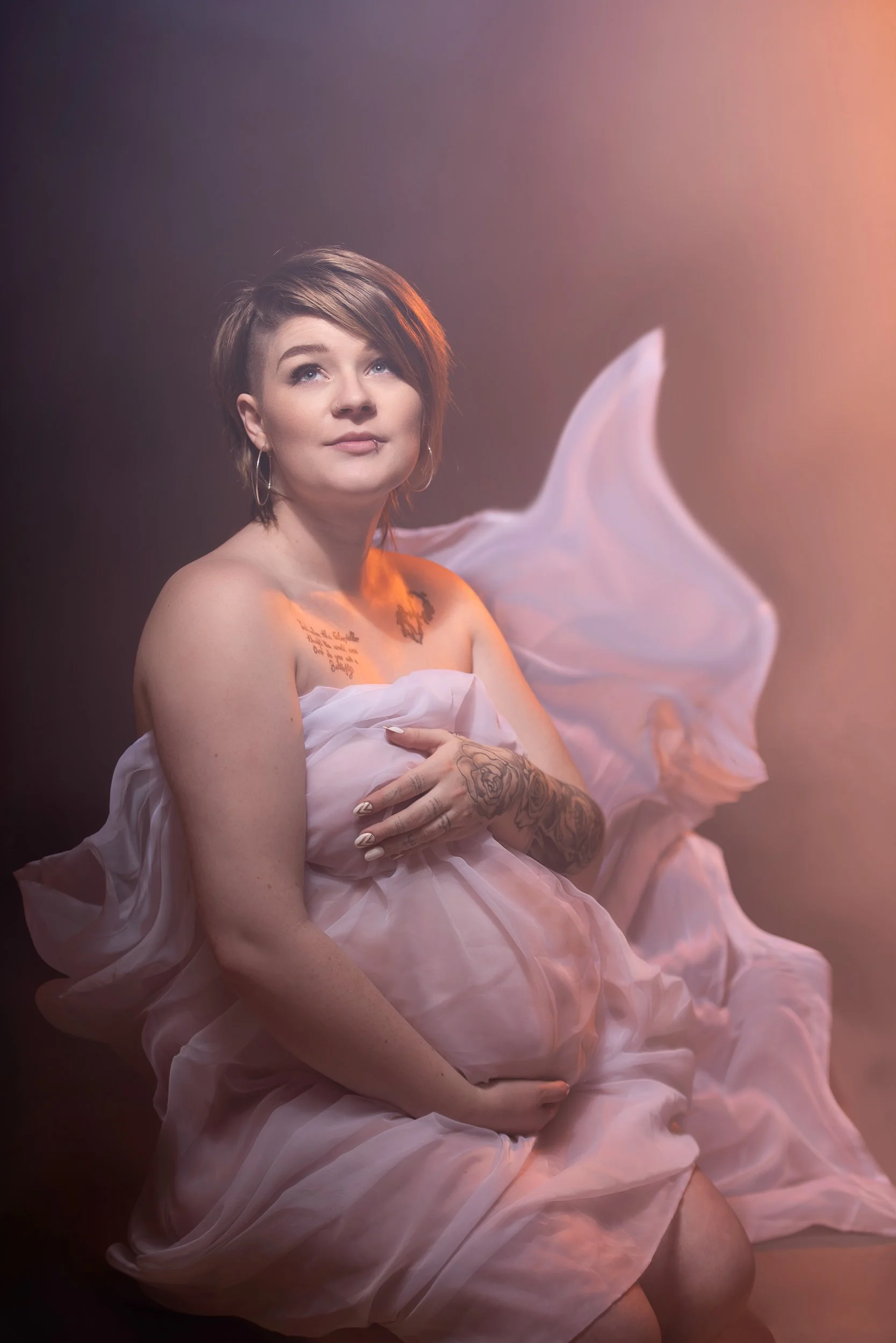 A pregnant woman with tattoos gently holding her belly, wrapped in flowing pink fabric with soft pink and purple lighting against a dark background.