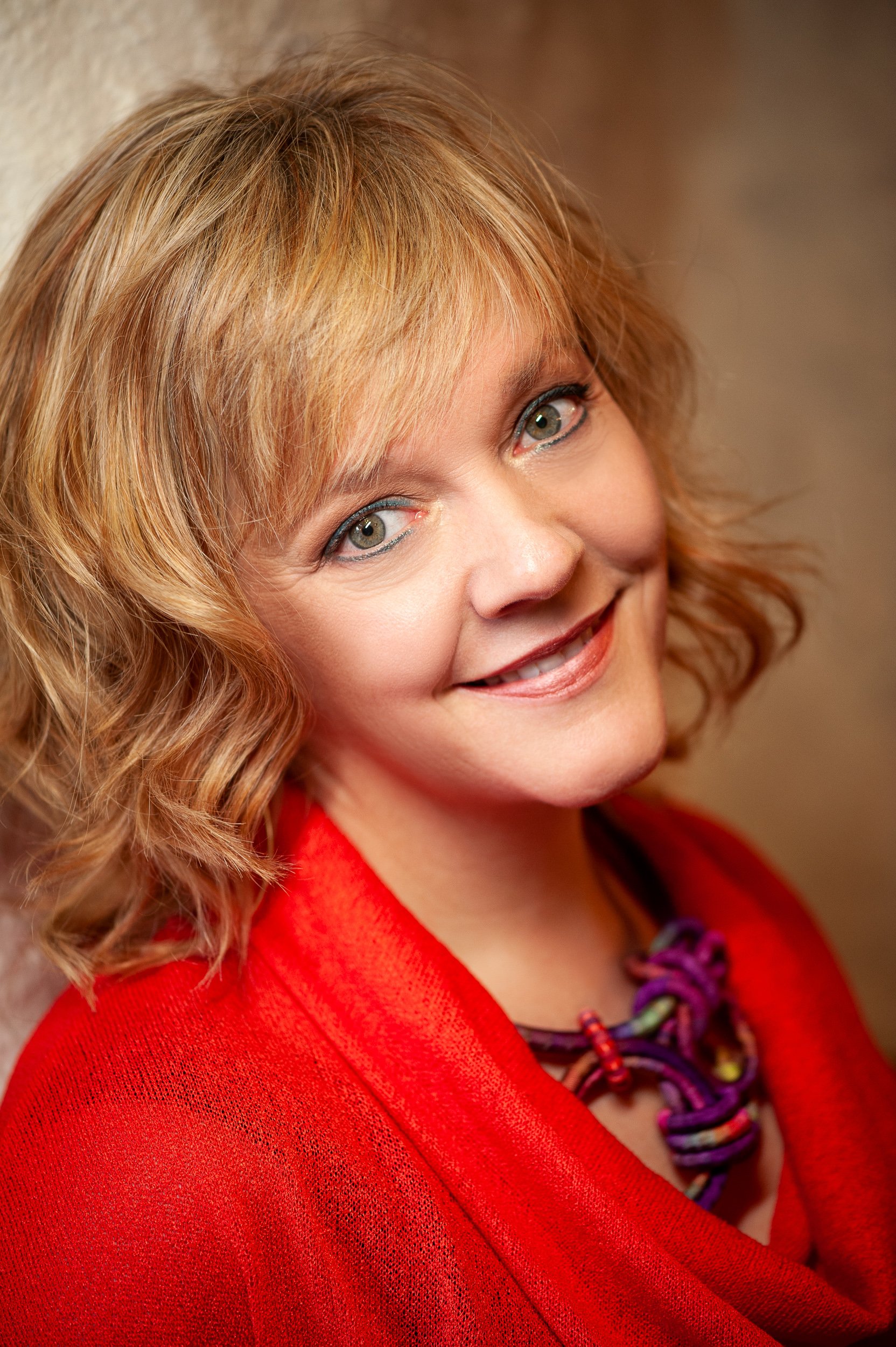 Portrait of a woman with shoulder-length blonde hair wearing a red top and colourful necklace, smiling naturally at the camera.