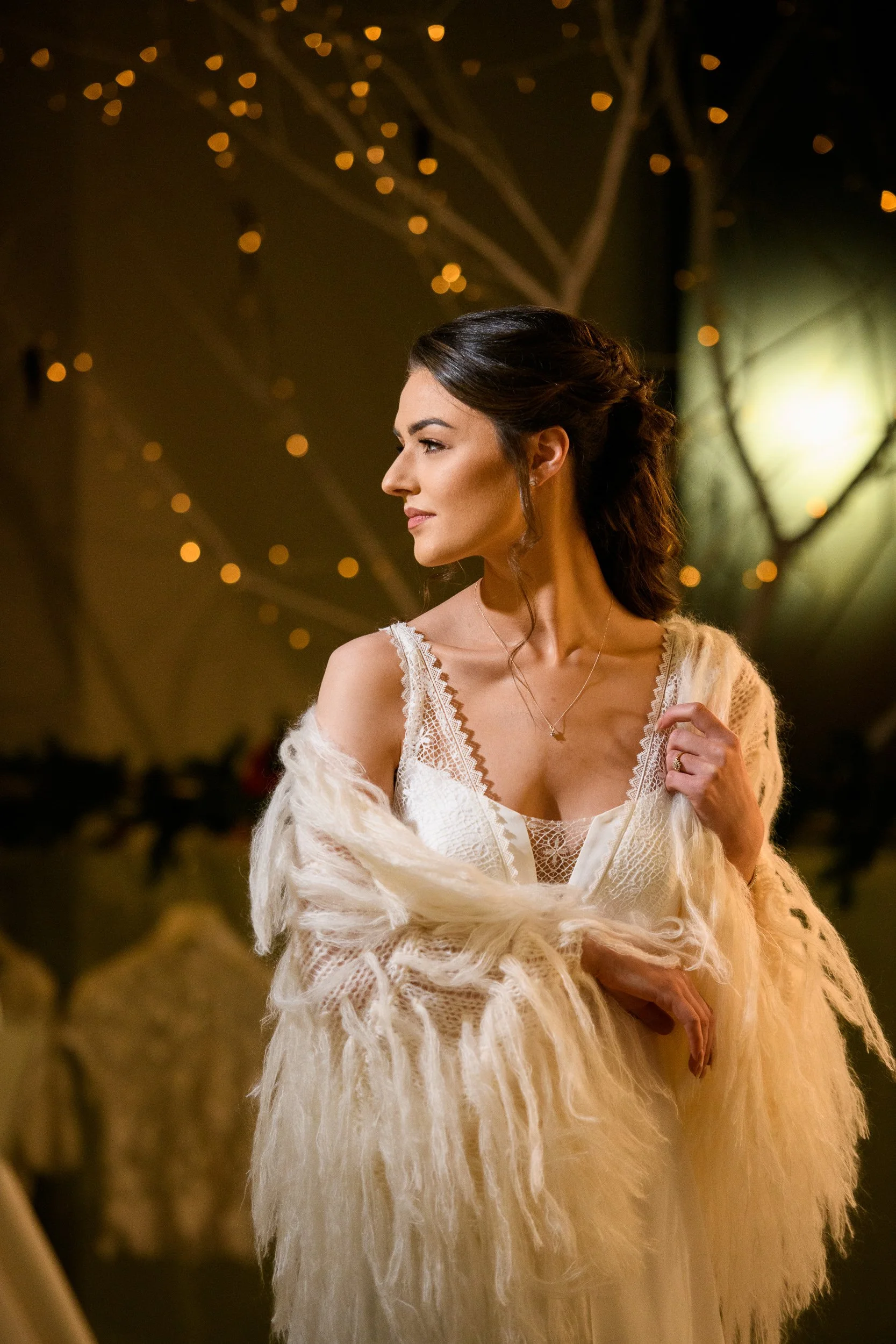 Elegant portrait of a woman in a lace white dress and soft shawl, standing in warm natural light indoors
