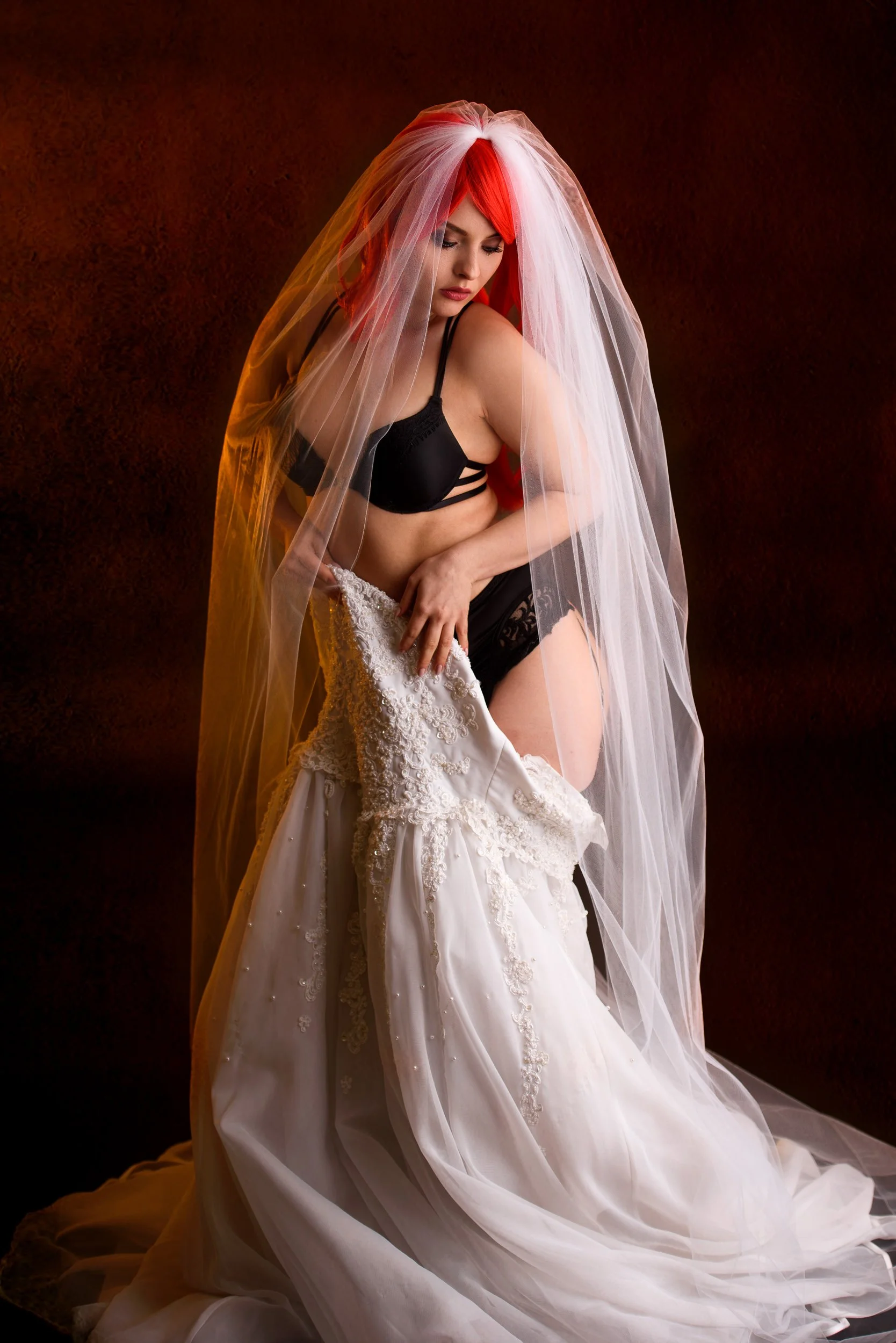 A woman with red and white hair wearing black lingerie and a veil, holding a wedding dress in a stylised, editorial setting.
