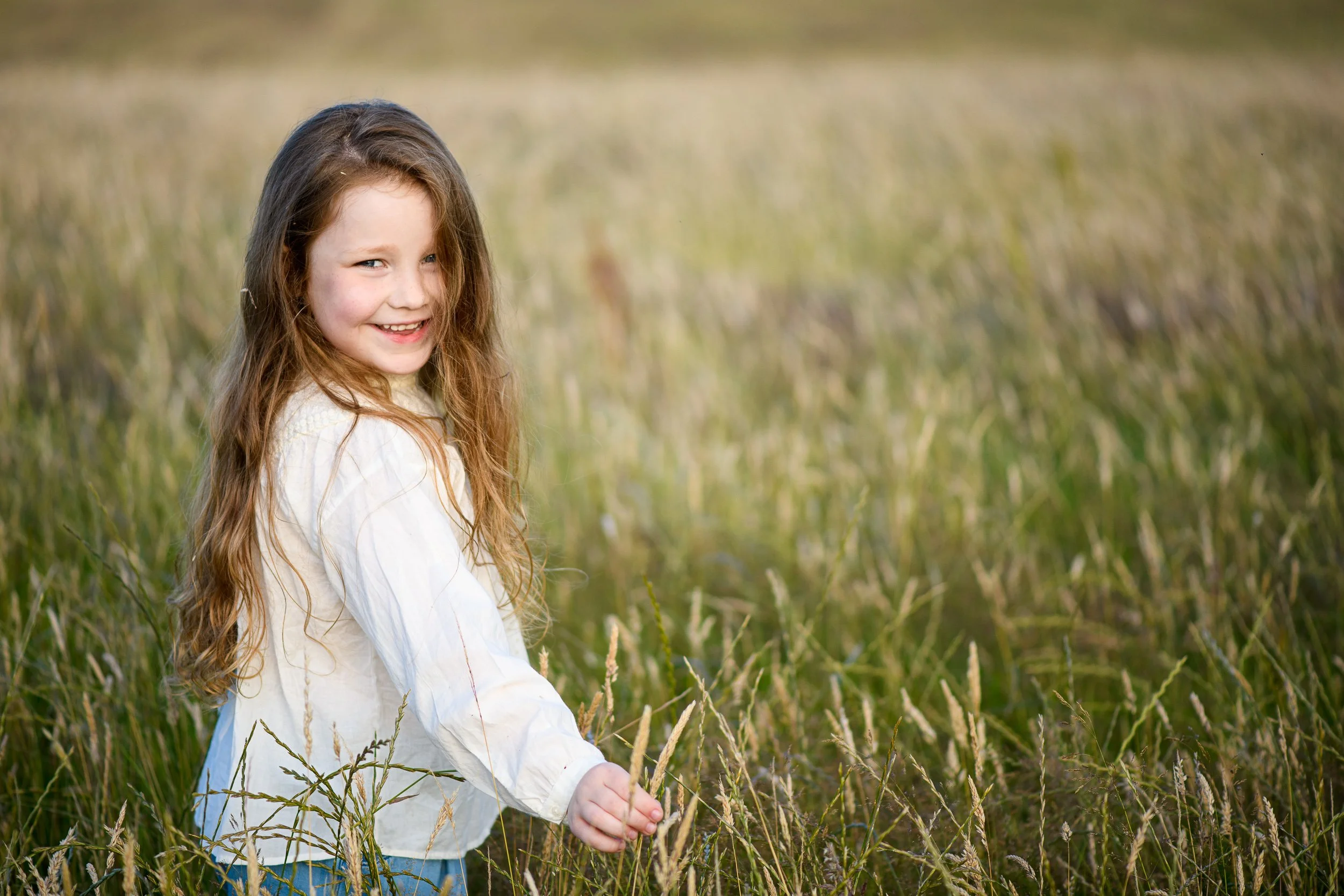A young girl with long red hair smiling while playing in a field of tall grass.
