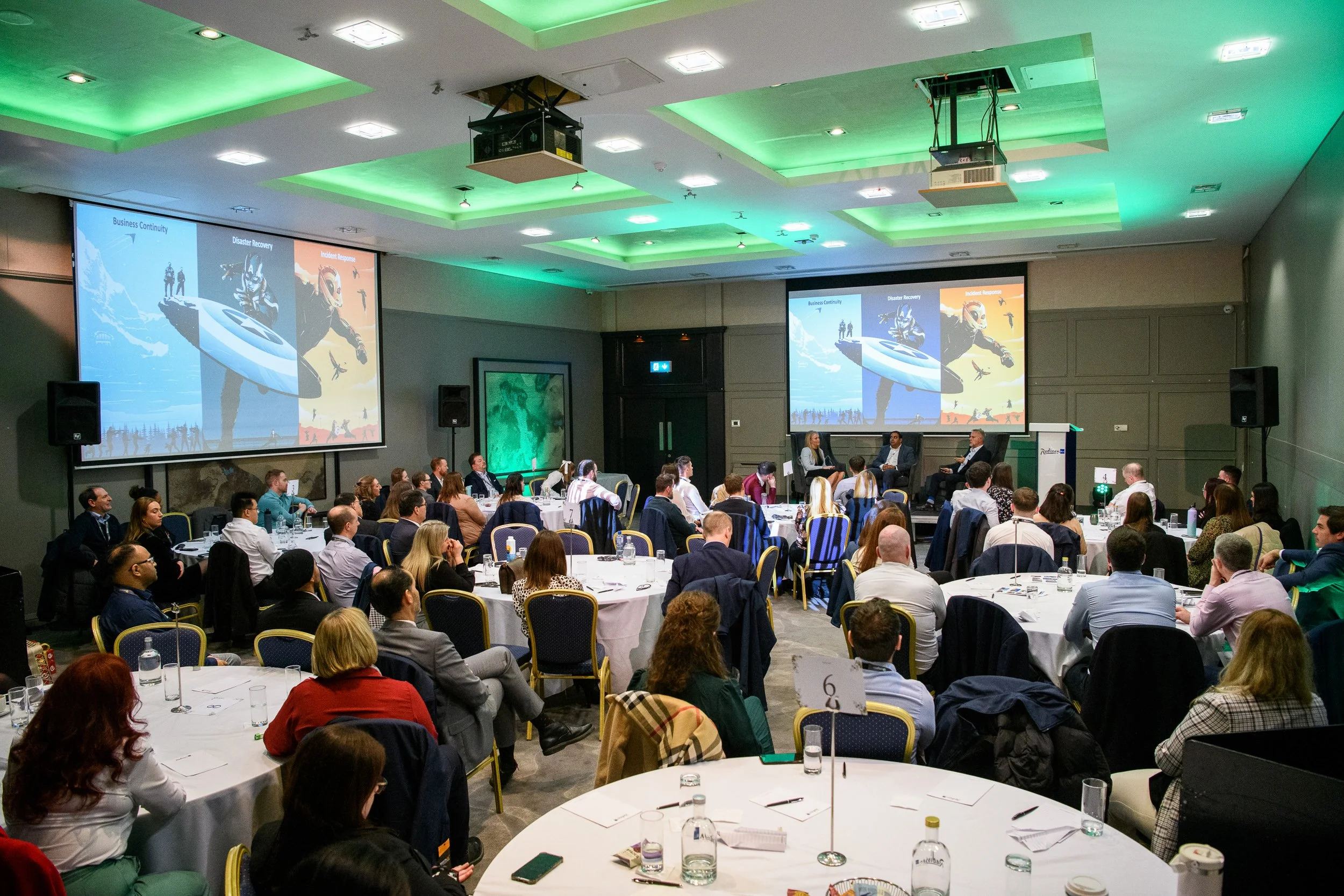 Conference audience seated at round tables facing a stage presentation in a large event venue.
