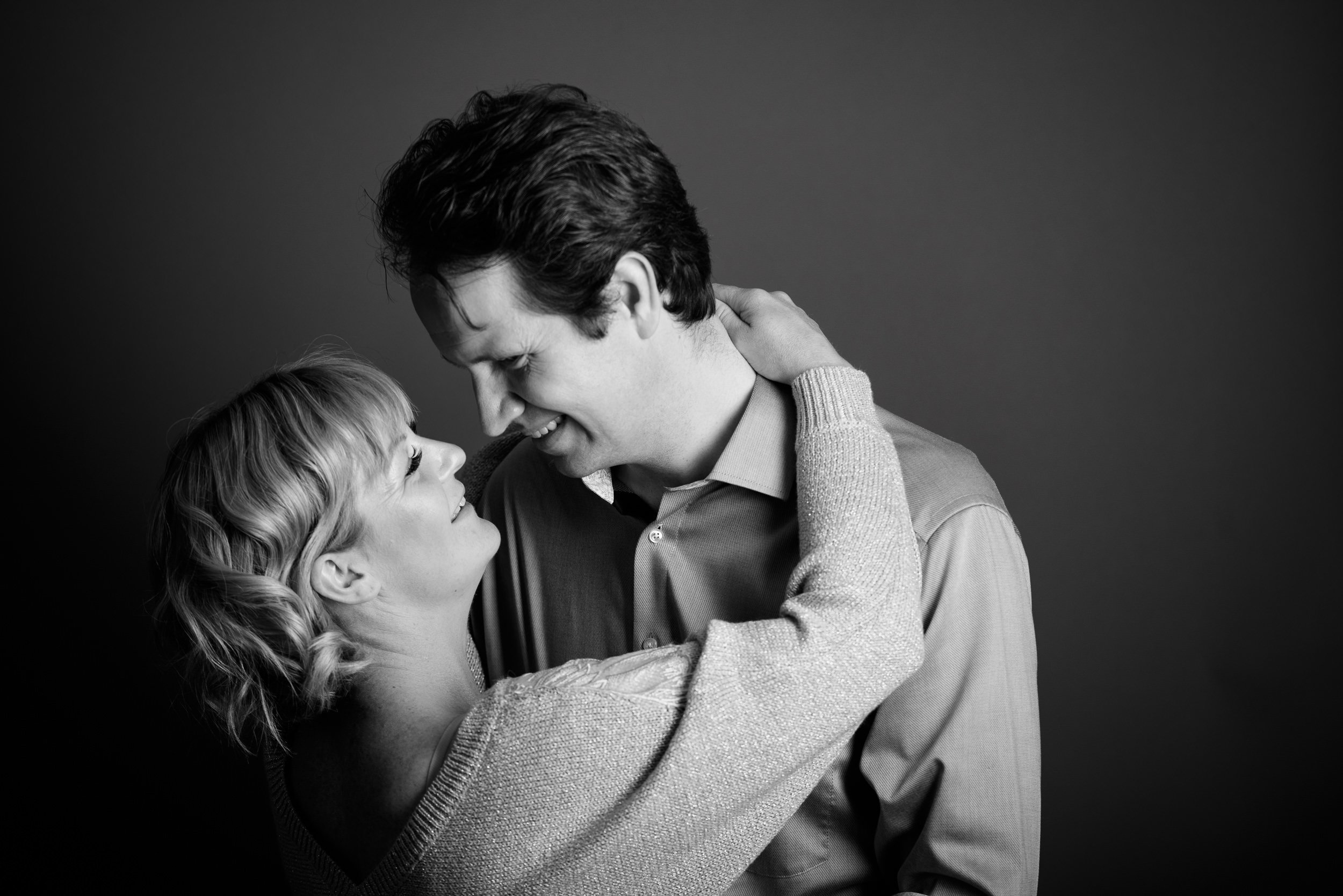A black-and-white photo of a woman and man looking into each other's eyes, smiling, with the woman's arm around the man's neck.