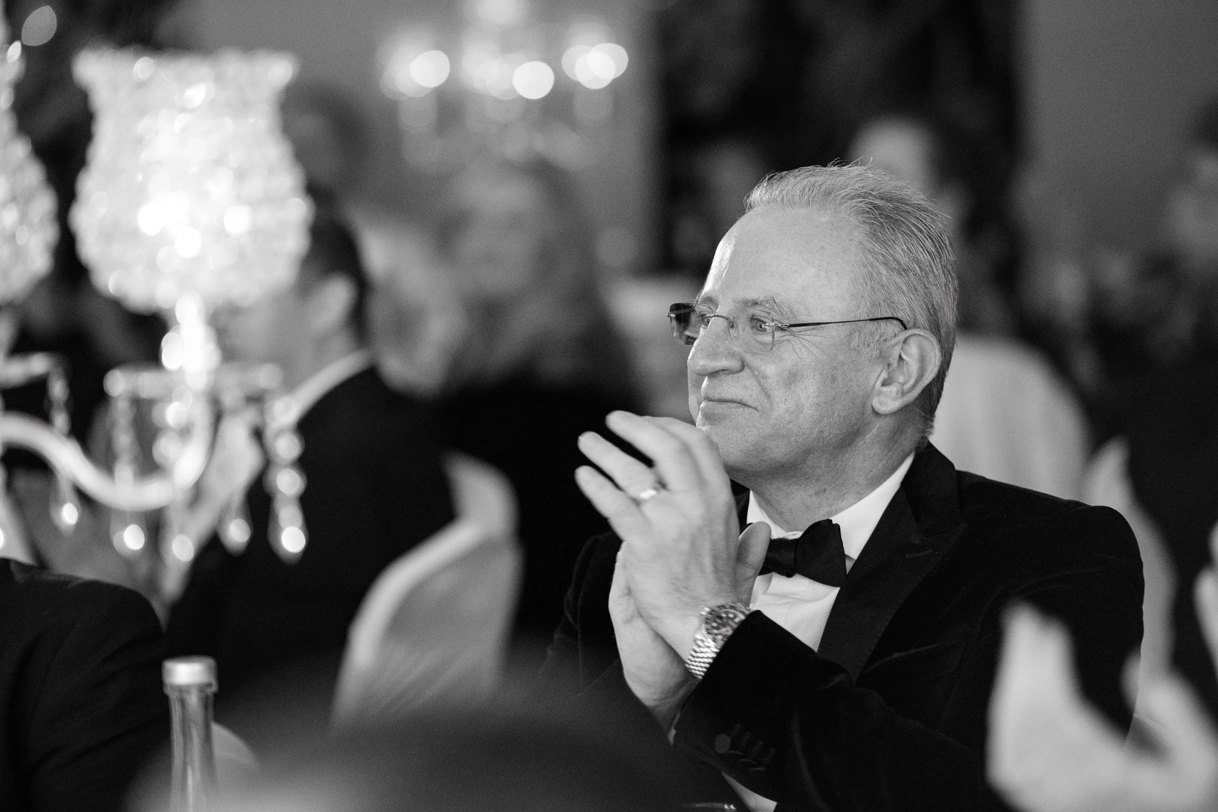 A man in formal tuxedo with glasses and a watch, sitting at a banquet, attentively listening at an elegant event.