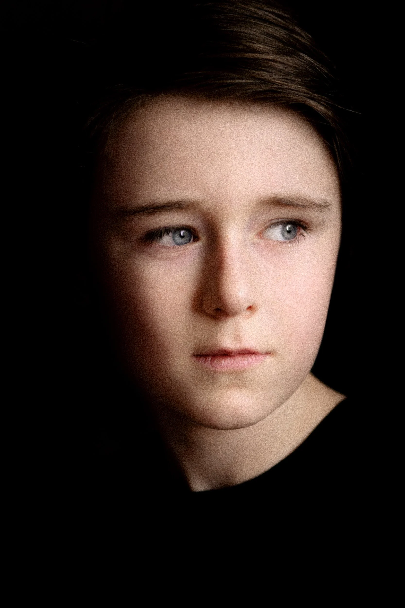 Ken Byrne Portrait Photography - Close-up portrait of a young girl with light skin, blue eyes, and short brown hair, looking off to the side against a black background.
