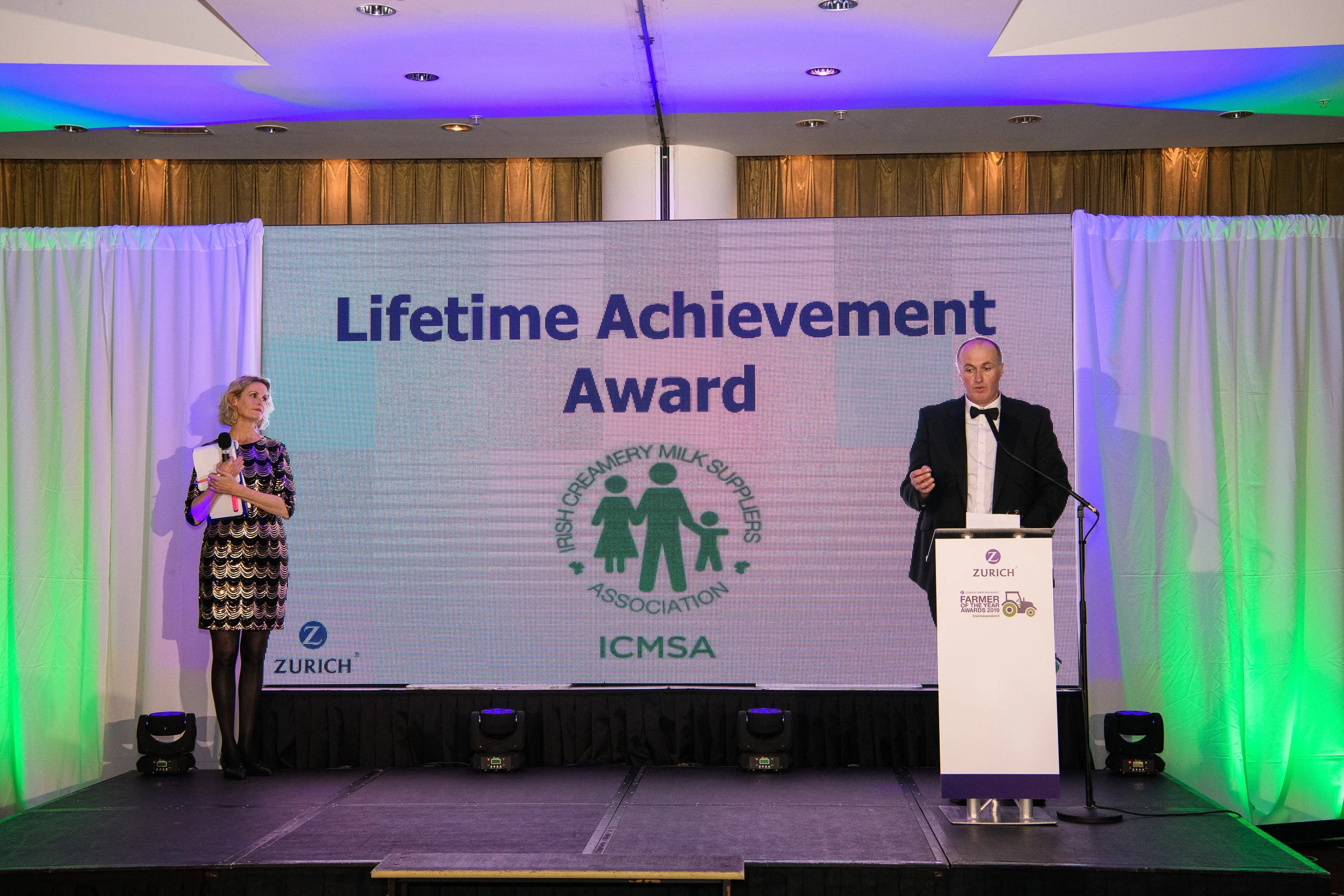 A man in a tuxedo speaks at a podium on stage, with a woman holding a microphone and papers standing to his left, at the 'Lifetime Achievement Award' ceremony for the Pastry, Dairy, Milk Suppliers Association, with a large screen behind them displayi