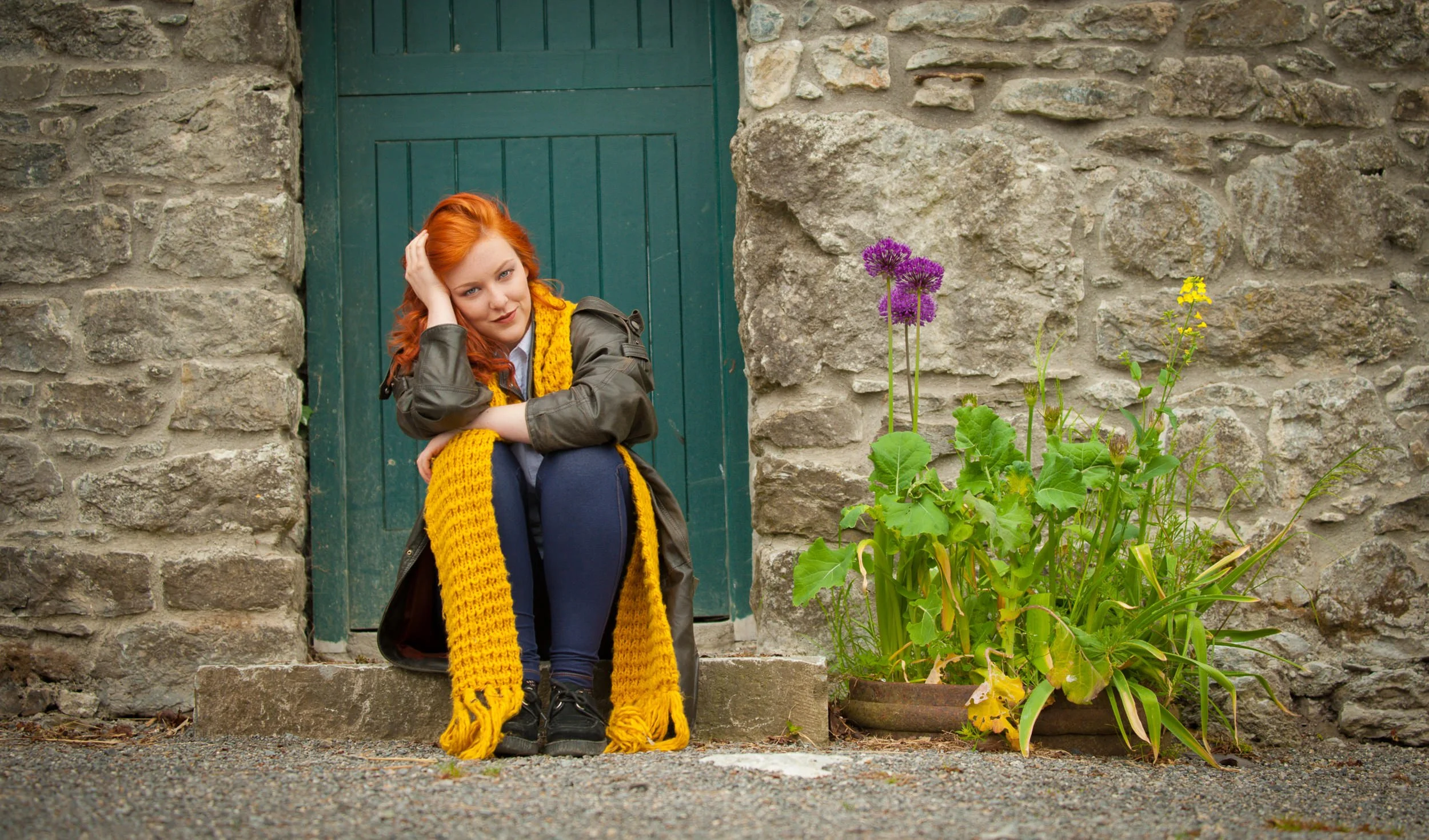 Outdoor portrait of a red-haired woman sitting by a green door with flowers and textured stone backdrop
