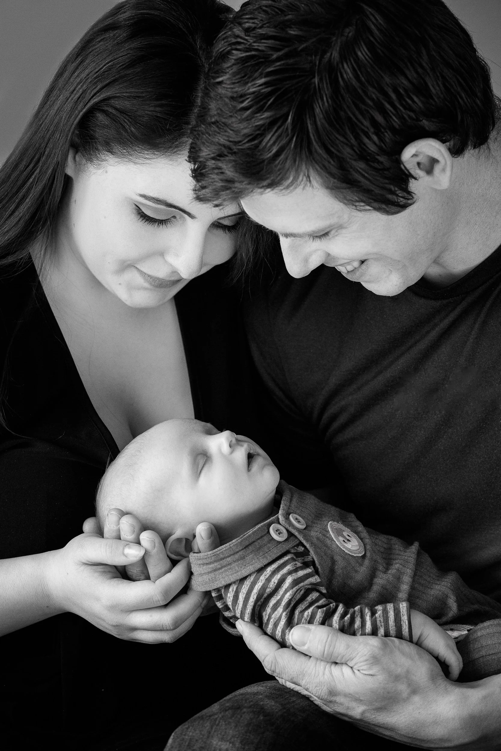 Black and white portrait of parents holding their sleeping baby, both looking at the child with affection.
