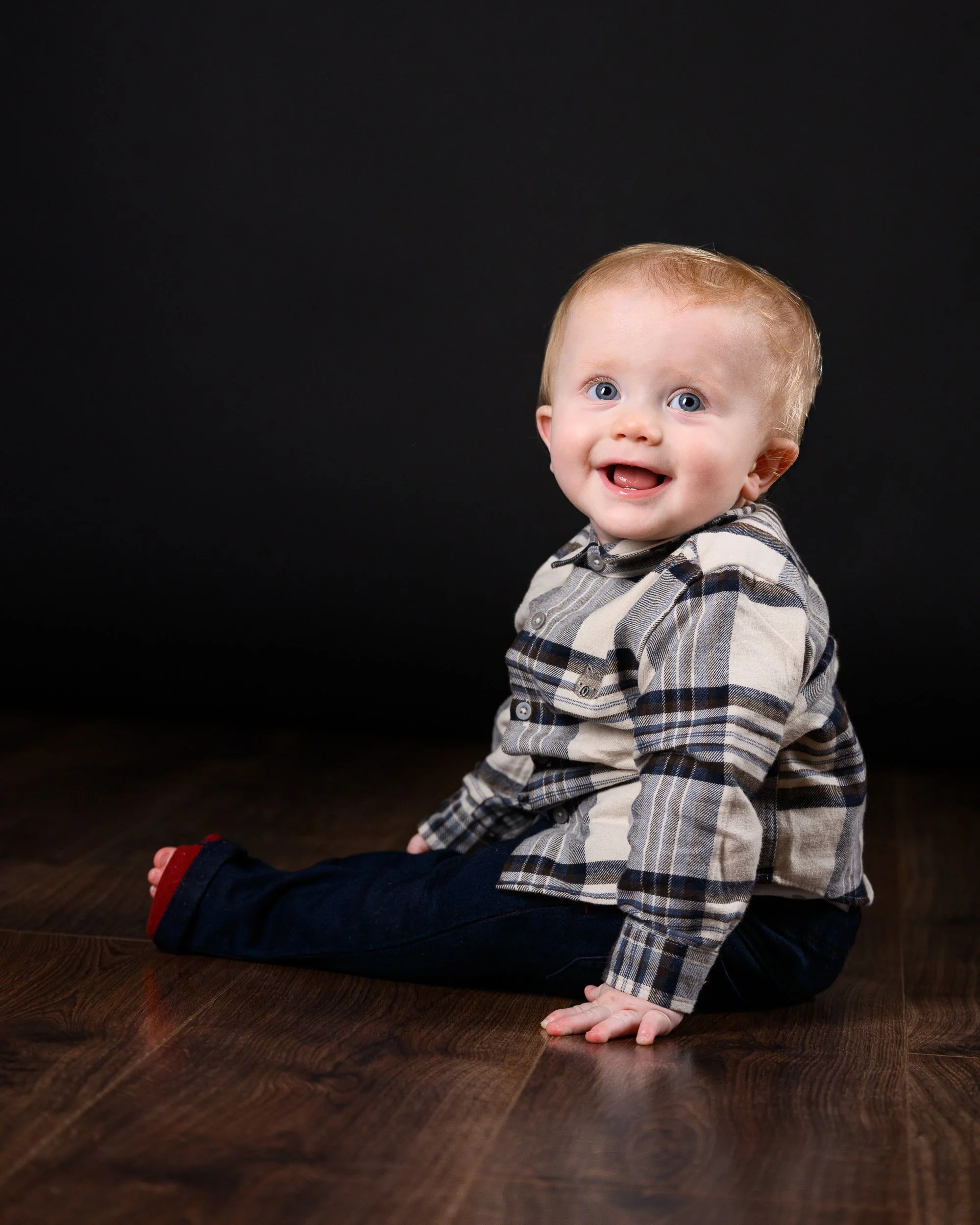 A baby with blue eyes sitting on a wooden floor, smiling against a dark background.
